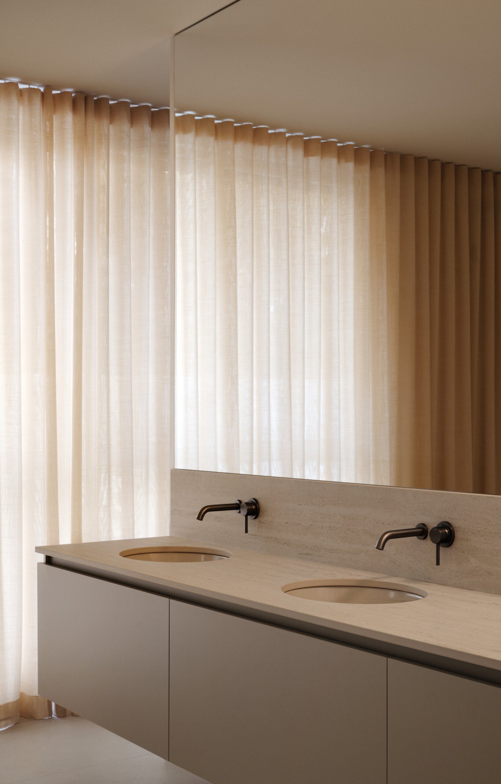 The primary bathroom is a quiet retreat, designed for comfort and calm. Sheer curtains filter soft light across a minimalist floating vanity with twin basins and a pale stone countertop. Muted metal fixtures add subtle contrast, while the mirrored above makes the room feel larger. With soothing tones, refined details, and no visual clutter, the ensuite feels spacious, tranquil, and perfectly suited for slow mornings. Two walk-in closets complete the suite with functional elegance.