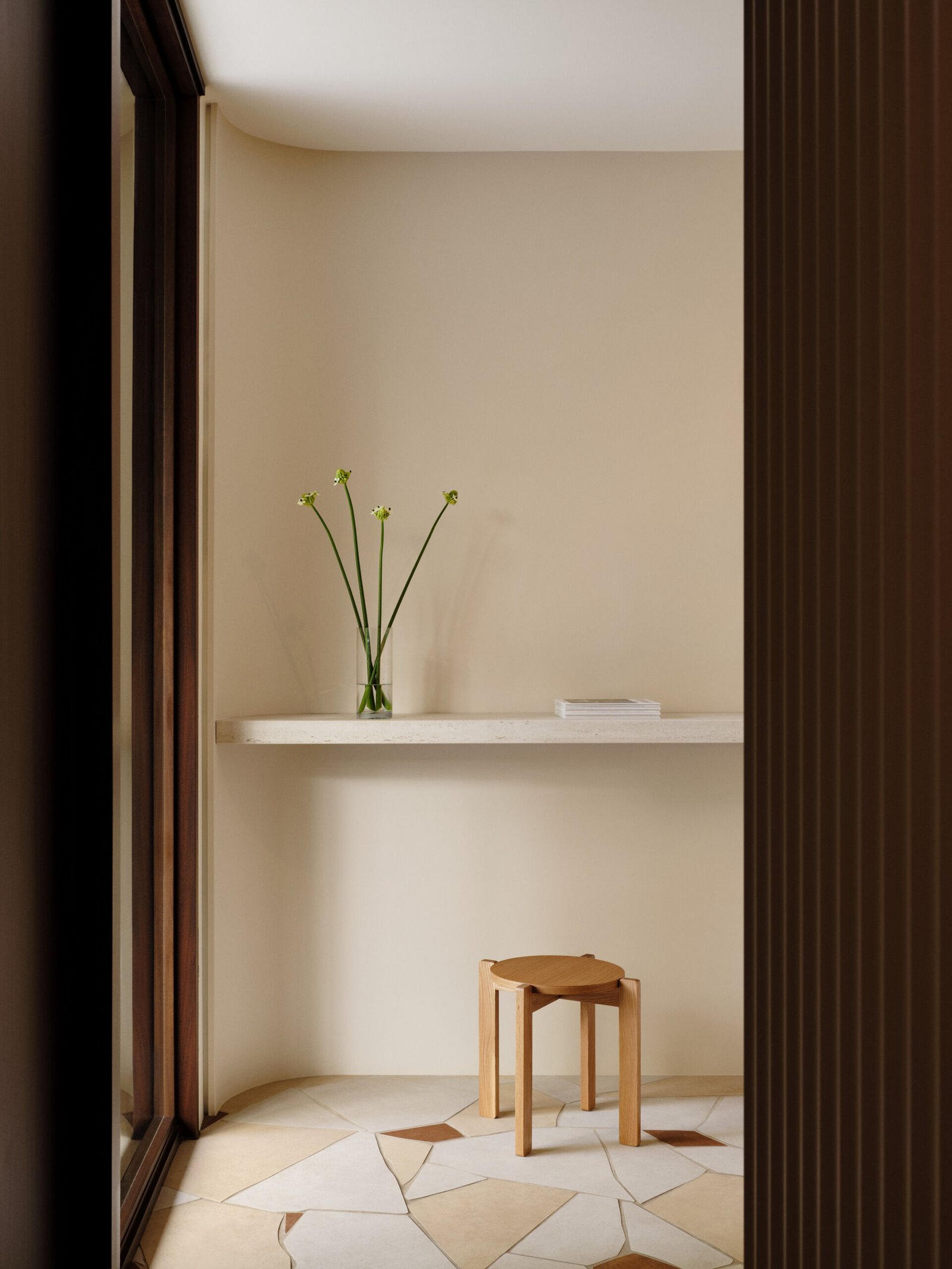 The entryway offers a quiet study in restraint and beauty. A simple floating shelf in pale stone runs along a softly curved wall, echoing the gentle geometry found throughout the home. The styling is minimal yet poetic—a slender glass vase with delicate green-stemmed blooms brings life to the space without disturbing its calm.Below, the floor steals a quiet moment of attention. Laid in an opus incertum pattern, the ceramic tiles resemble irregular shards of natural slate, with tones of cream, sand, and terracotta that add depth and warmth. A low, sculptural stool in natural oak sits alone, its softly rounded edges mirroring the architectural gestures found throughout the house.This small space captures the essence of the entire home: quiet elegance, natural light, and a deep respect for materiality.