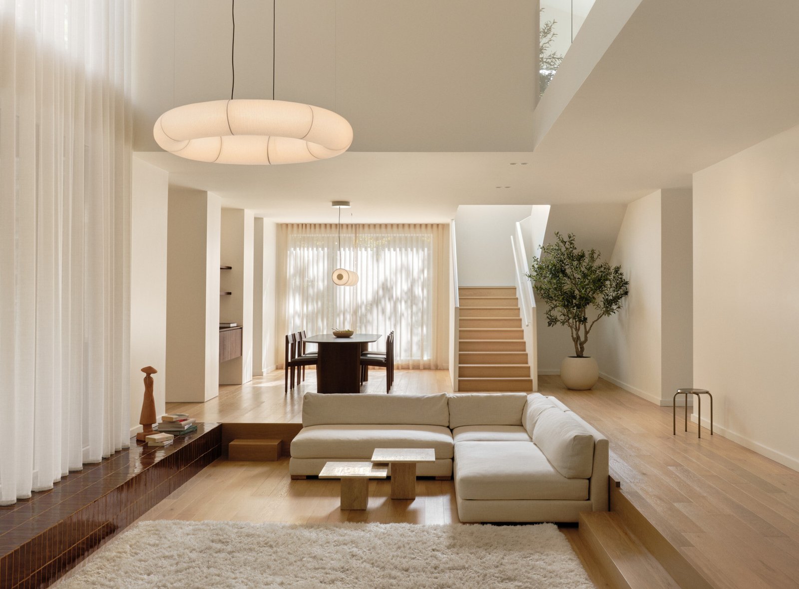 Slightly sunken and wrapped in warmth, the living room feels carved from the landscape itself. A linear fireplace positioned on a glazed terracotta tile platform creates an inviting, grounded space centered around a plush ivory rug. Soft curves in the white plaster echo the home's rounded details, while pale built-in sofas and a black leather lounge chair add contrast and calm. A sheer curtain highlights the double-height ceiling, and a glowing washi paper pendant adds warmth.