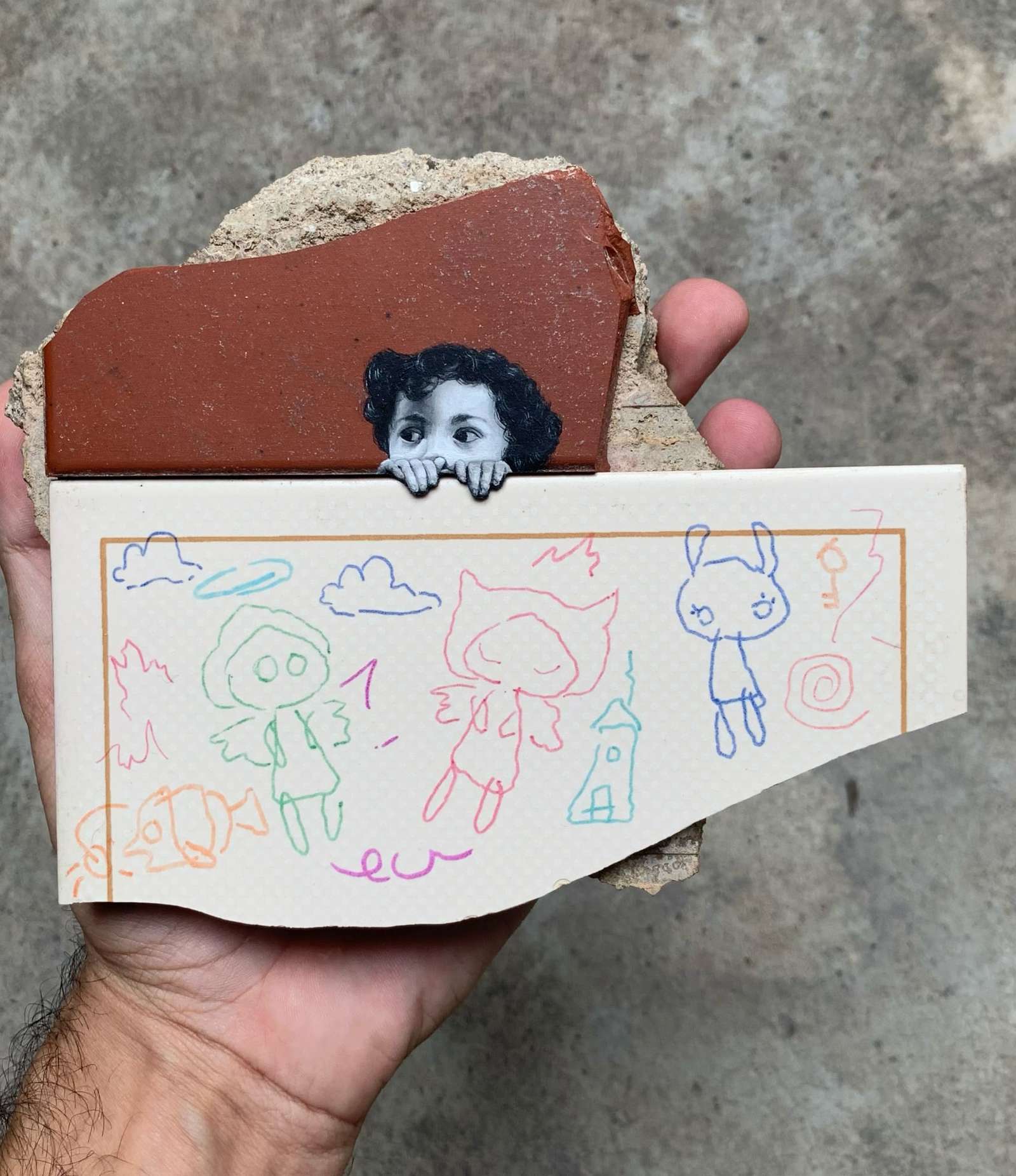 a hand holding an architectural fragment with a painting of a child peeking over a tile with colorful line drawings