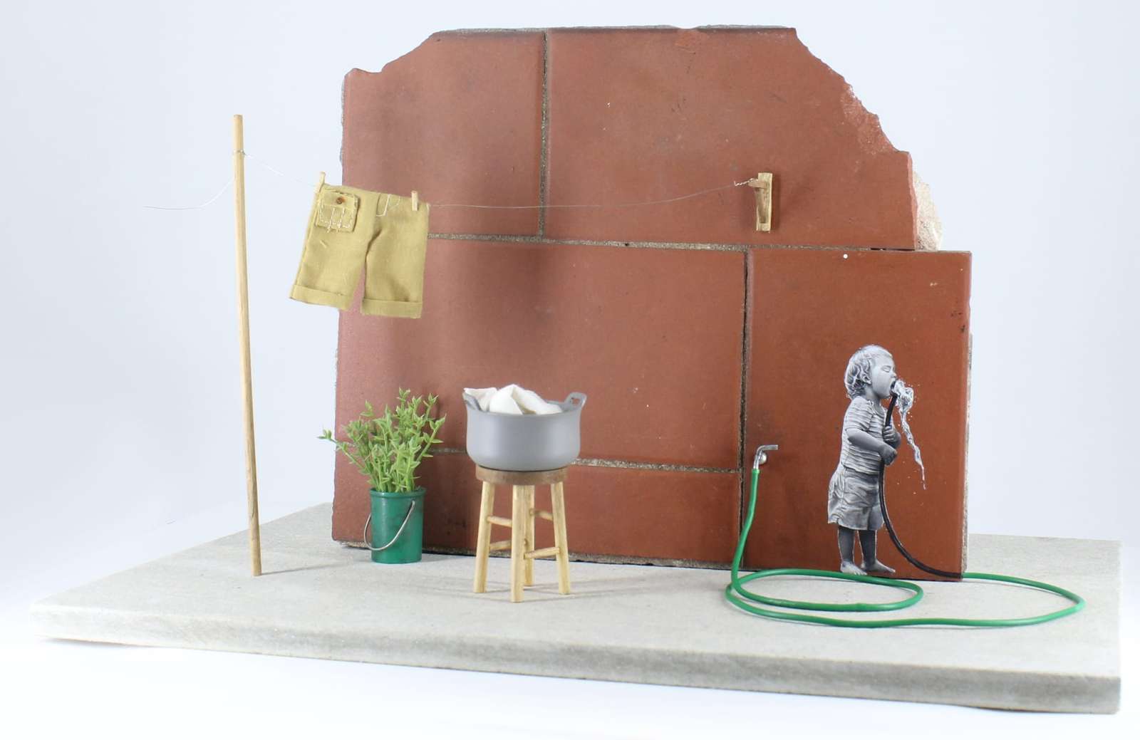 a child drinking from a green hose painted on tile with a wash bucket and laundry on the line nearby