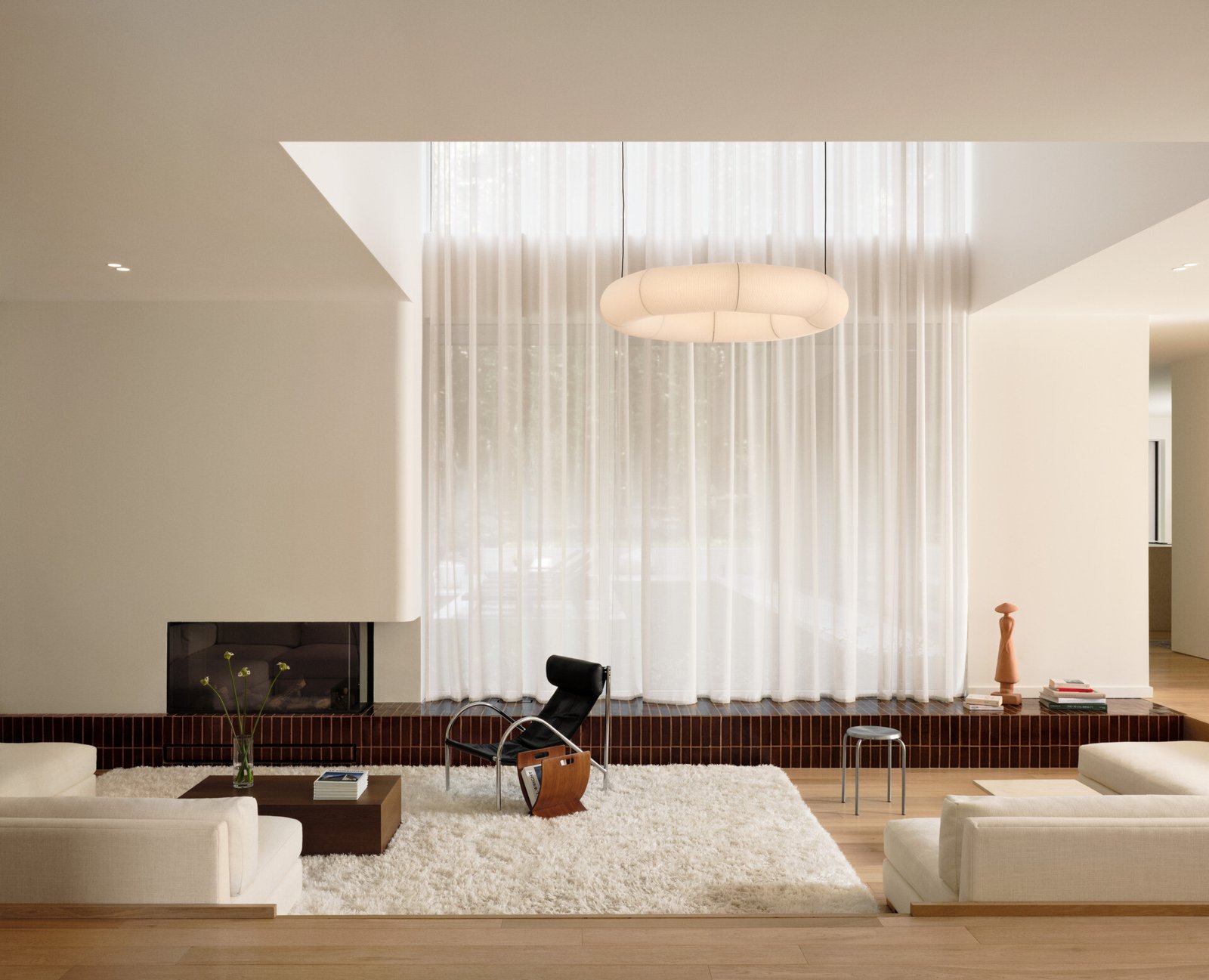 This sunken living room is both sculptural and serene. It creates an inviting, grounded space centered around a plush ivory rug and a terracotta tile platform. Rich in tone and laid with clean precision, the tile provides a grounding base for both the fireplace and the window wall.Soft curves in the white plaster echo the home's rounded details, while pale sofas and a black leather lounge chair add contrast and calm. A sheer curtain highlights the double-height ceiling, and a glowing washi paper pendant adds warmth.