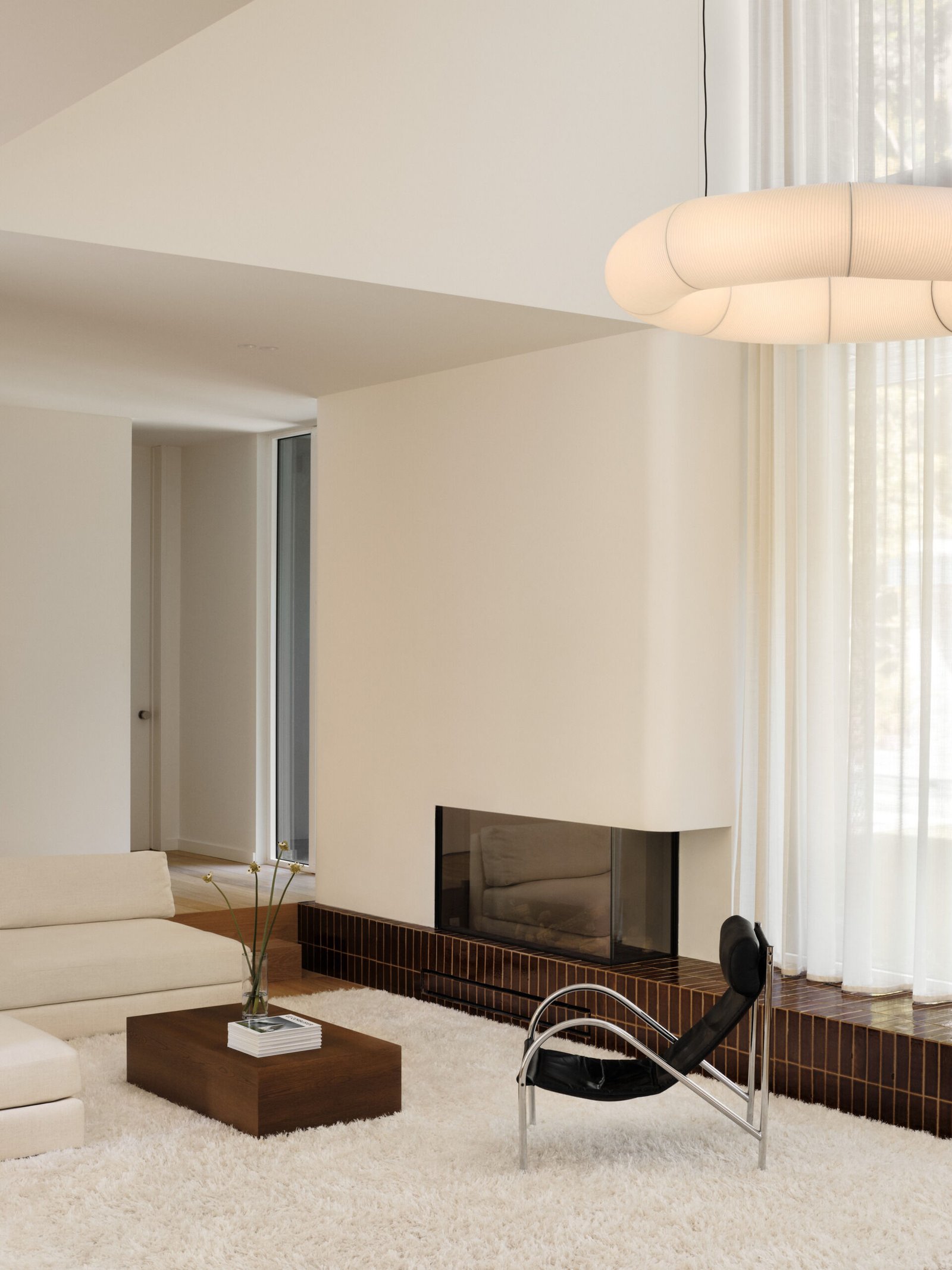 This sunken living room is both sculptural and serene. It creates an inviting, grounded space centered around a plush ivory rug and a terracotta tile platform. Rich in tone and laid with clean precision, the tile provides a grounding base for both the fireplace and the window wall.Soft curves in the white plaster echo the home's rounded details, while pale sofas and a black leather lounge chair add contrast and calm. A sheer curtain highlights the double-height ceiling, and a glowing washi paper pendant adds warmth.