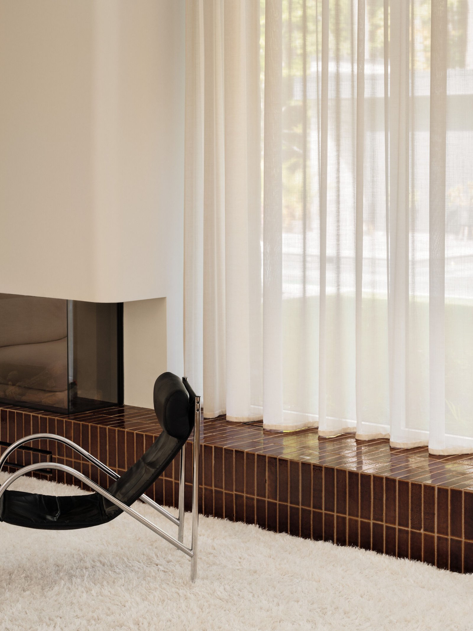 From above, the living room reveals a balanced composition of form and texture. A black leather lounge chair and dark wood coffee table sit on a plush white rug, creating a refined contrast. The fireplace, set into a rounded white plaster wall and framed in glossy terracotta tile, adds quiet depth while defining the space with a bold horizontal line.