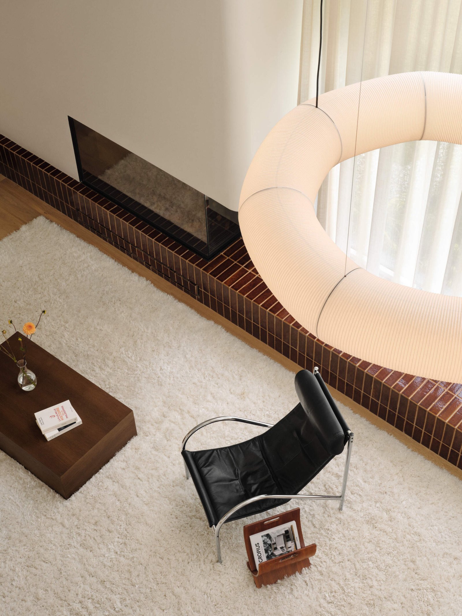 From above, the living room reveals a balanced composition of form and texture. A black leather lounge chair and dark wood coffee table sit on a plush white rug, creating a refined contrast. The fireplace, set into a rounded white plaster wall and framed in glossy terracotta tile, adds quiet depth while defining the space with a bold horizontal line.