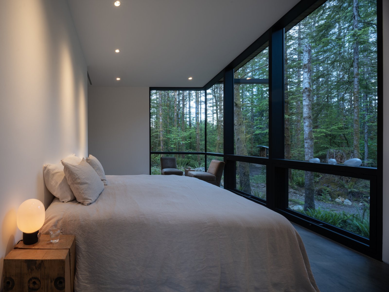 Surrounded by forest and softened by natural light, the bedroom offers a quiet, secluded retreat. Floor-to-ceiling windows frame the trees outside, providing both views and privacy. Simple furnishings and pale bedding keep the space understated, while a warm bedside light adds gentle illumination.