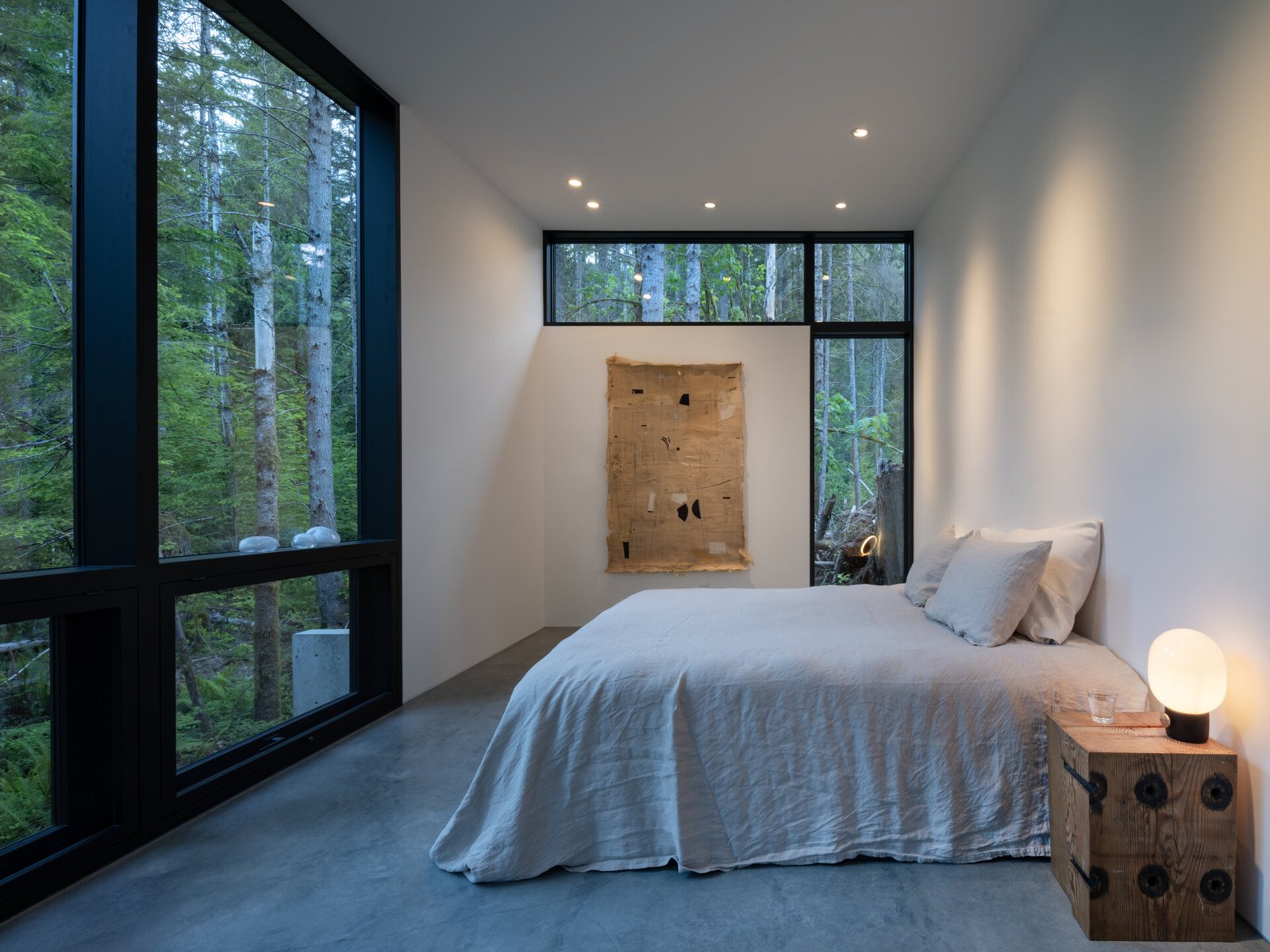 Surrounded by forest and softened by natural light, the bedroom offers a quiet, secluded retreat. Floor-to-ceiling windows frame the trees outside, providing both views and privacy. Simple furnishings and pale bedding keep the space understated, while a warm bedside light adds gentle illumination.