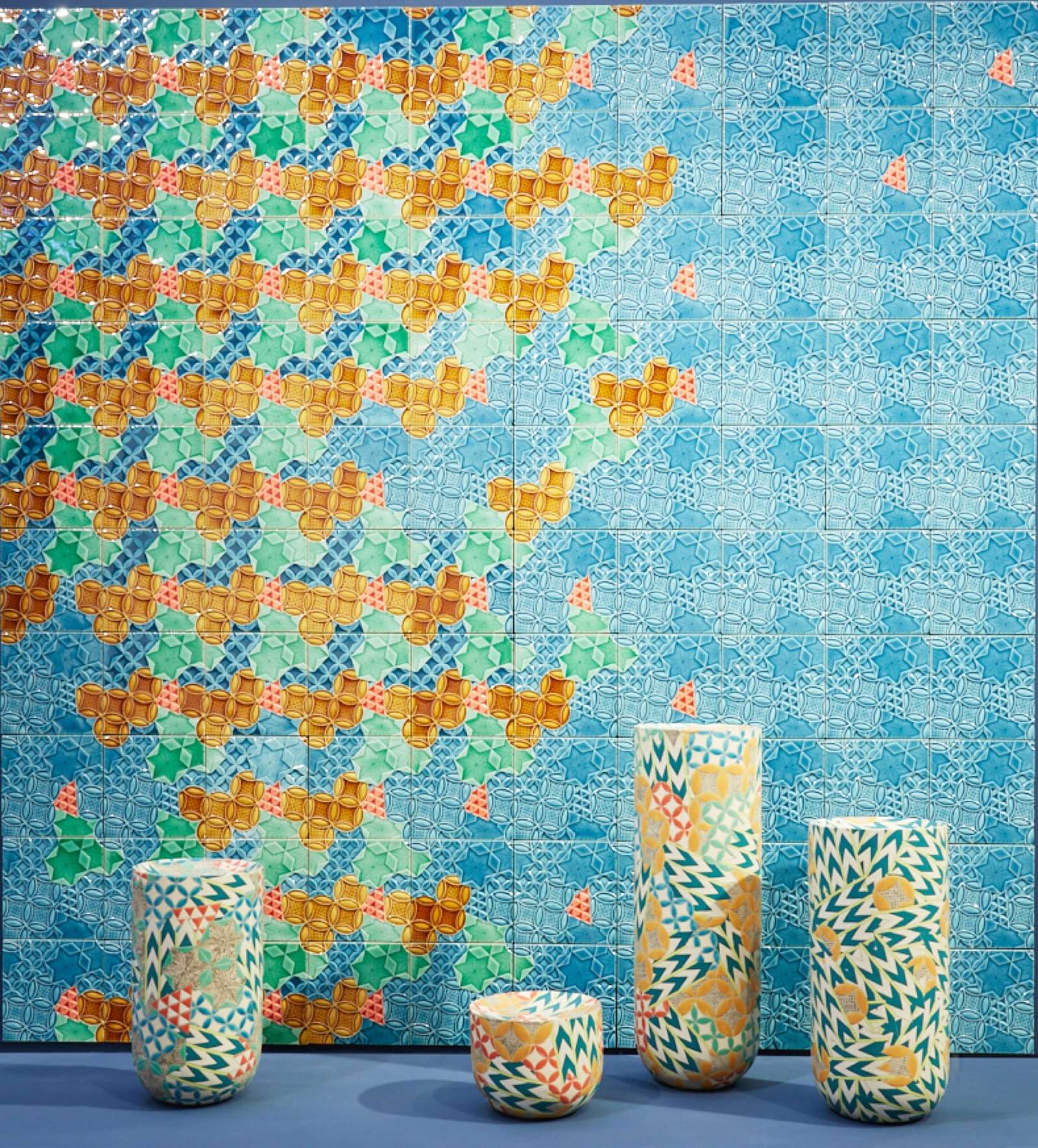 four colorful, geometrically patterned, cylindrical ceramic forms in front of a patterned tile backdrop