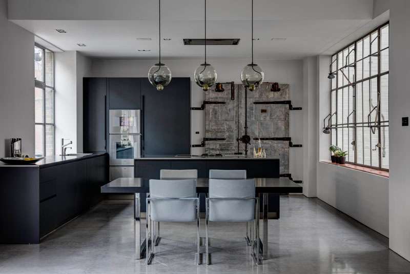 In the kitchen, a striking old industrial door has been incorporated into the design, tying the home back to its factory roots. Black cabinetry contrasts against the crisp white walls and polished concrete flooring, while the warm tones of wood and subtle industrial hues add depth. 