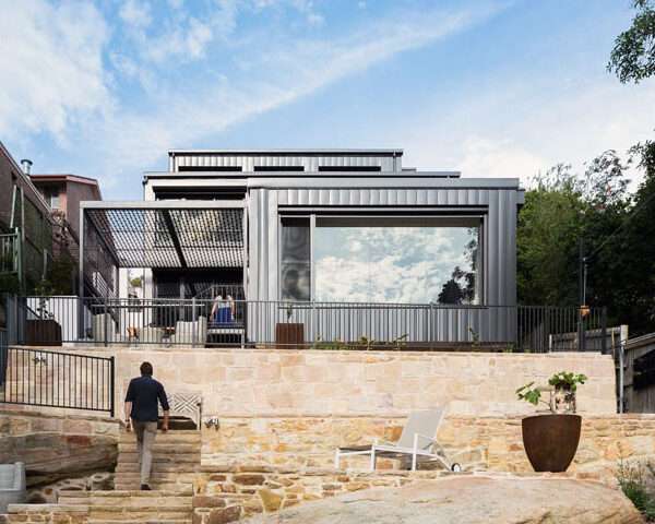 A contemporary home rises along a steep slope and blends seamlessly with the surrounding bushland. Built with fire-resistant materials and steel shutters for protection, the house balances safety with style, featuring multi-level living, expansive picture windows, and a strong connection between indoors and outdoors.