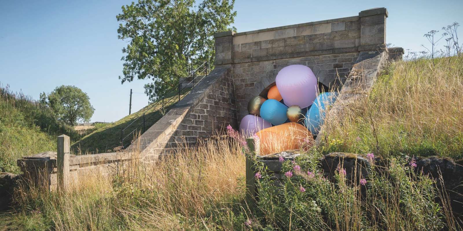 a large-scale installation by Steve Messam under an old stone bridge of numerous pastel-colored, bulbous forms stuck under an archway