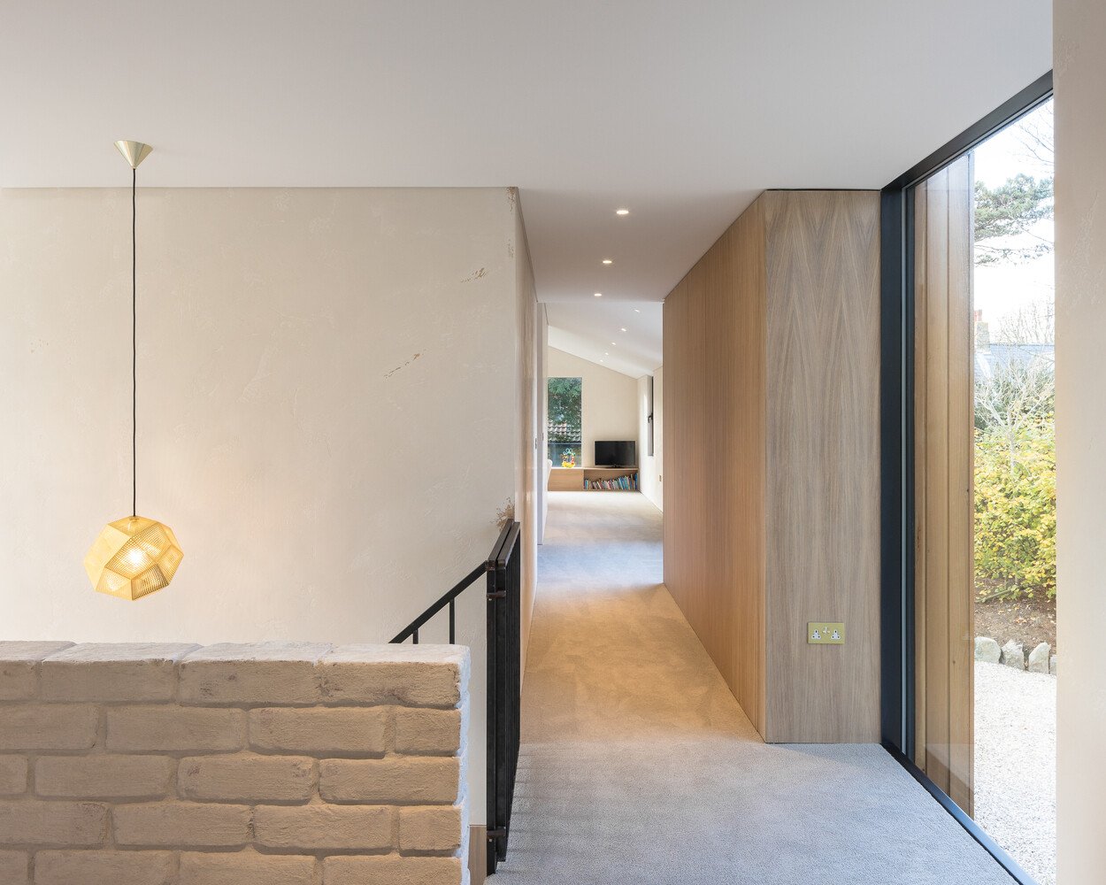 A bespoke raw steel staircase connects the two levels, sitting beside a wall of locally reclaimed brick coated in lime slurry. The contrast between metal and masonry adds texture to the journey upward, while soft light filters through from the double-height glazing that links both wings of the house.