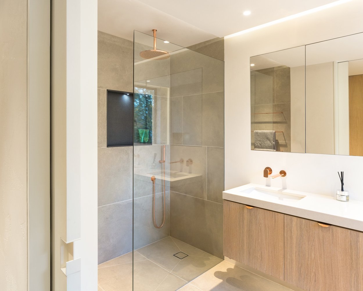 In the ensuite bathroom, a walk-in shower with a rainfall head and concealed lighting creates a spa-like calm. Subtle uplighting above the mirror enhances the gentle tones of the materials, while a skylight above the toilet area allows natural light to pour in, making even the smallest corner feel open.