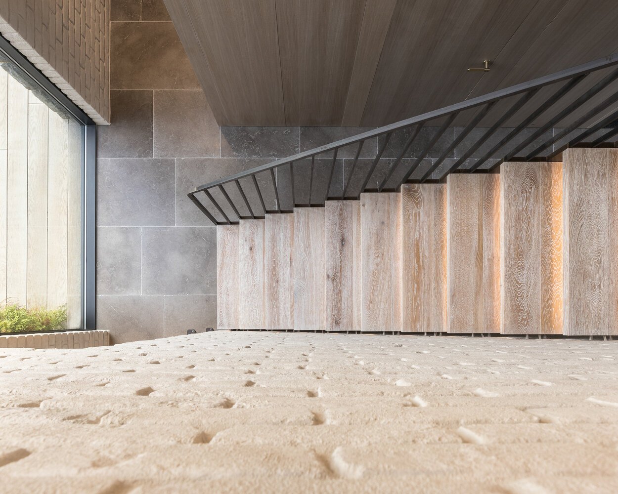 A bespoke raw steel staircase connects the two levels, sitting beside a wall of locally reclaimed brick coated in lime slurry. The contrast between metal and masonry adds texture to the journey upward, while soft light filters through from the double-height glazing that links both wings of the house.