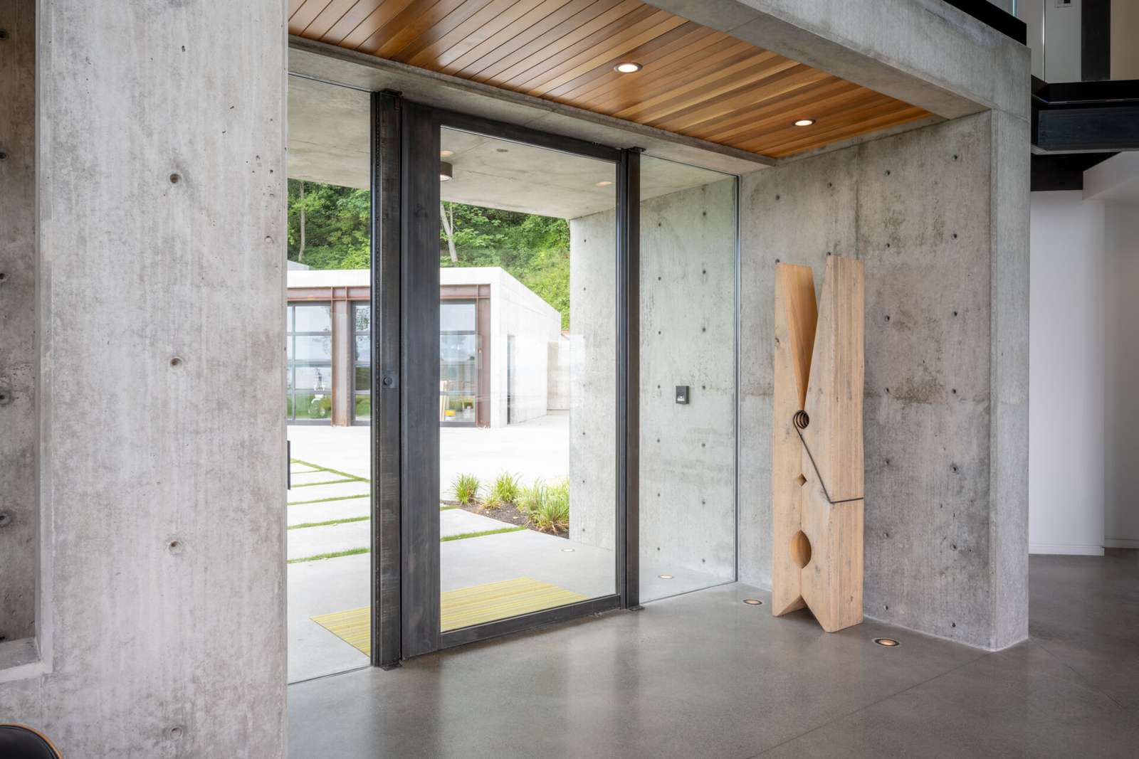 The entry is defined by exposed architectural concrete, immediately establishing the home’s material language. Above the entry sits a green roof, which becomes visible from inside the house. This moment connects the exterior landscape to the interior circulation, while a pivoting glass door welcomes visitors.