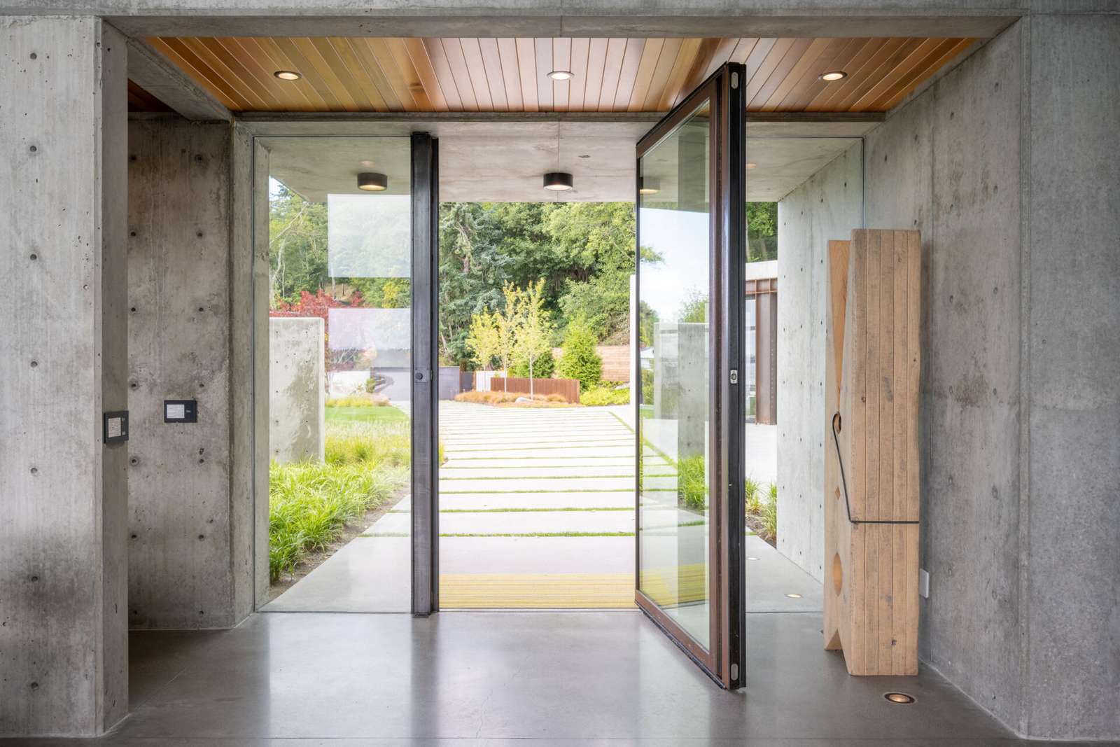 The entry is defined by exposed architectural concrete, immediately establishing the home’s material language. Above the entry sits a green roof, which becomes visible from inside the house. This moment connects the exterior landscape to the interior circulation, while a pivoting glass door welcomes visitors.