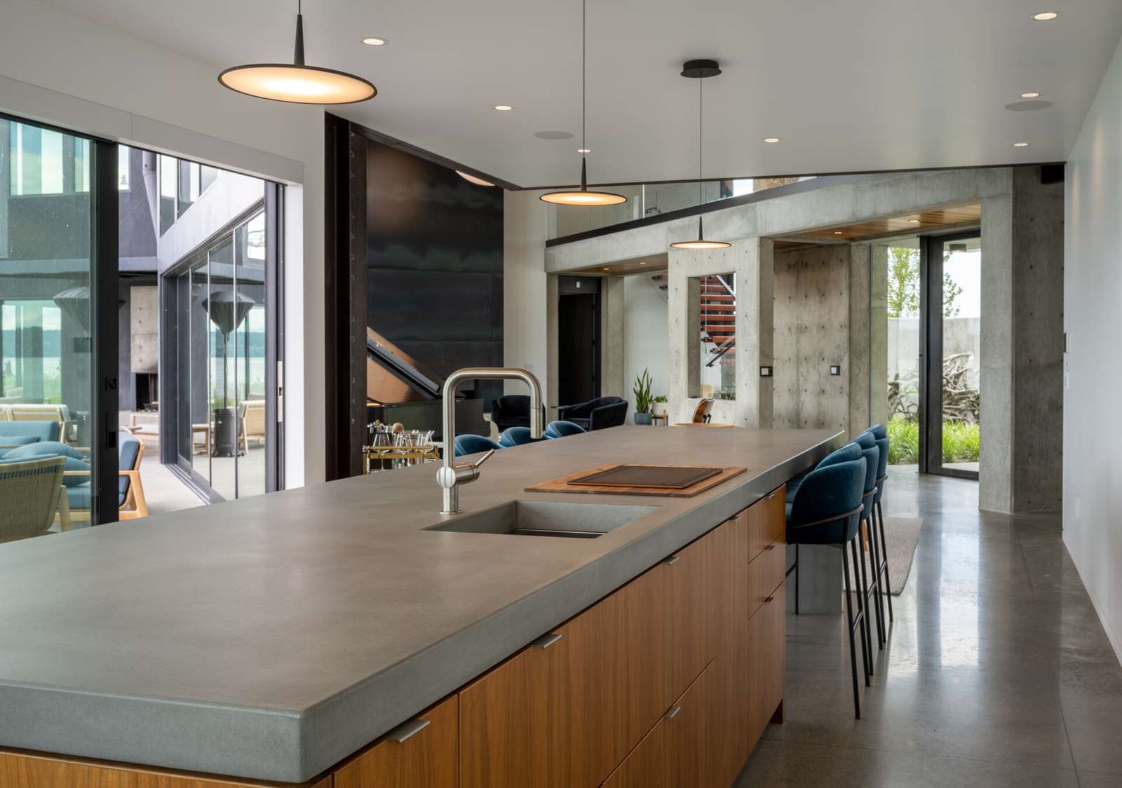 The kitchen has a long concrete island anchors the space, providing room for everyday use and casual gathering. Minimalist white cabinetry is paired with warm wood base units, keeping the palette restrained while adding subtle contrast. Floor to ceiling windows line the perimeter, drawing in natural light and uninterrupted water views.
