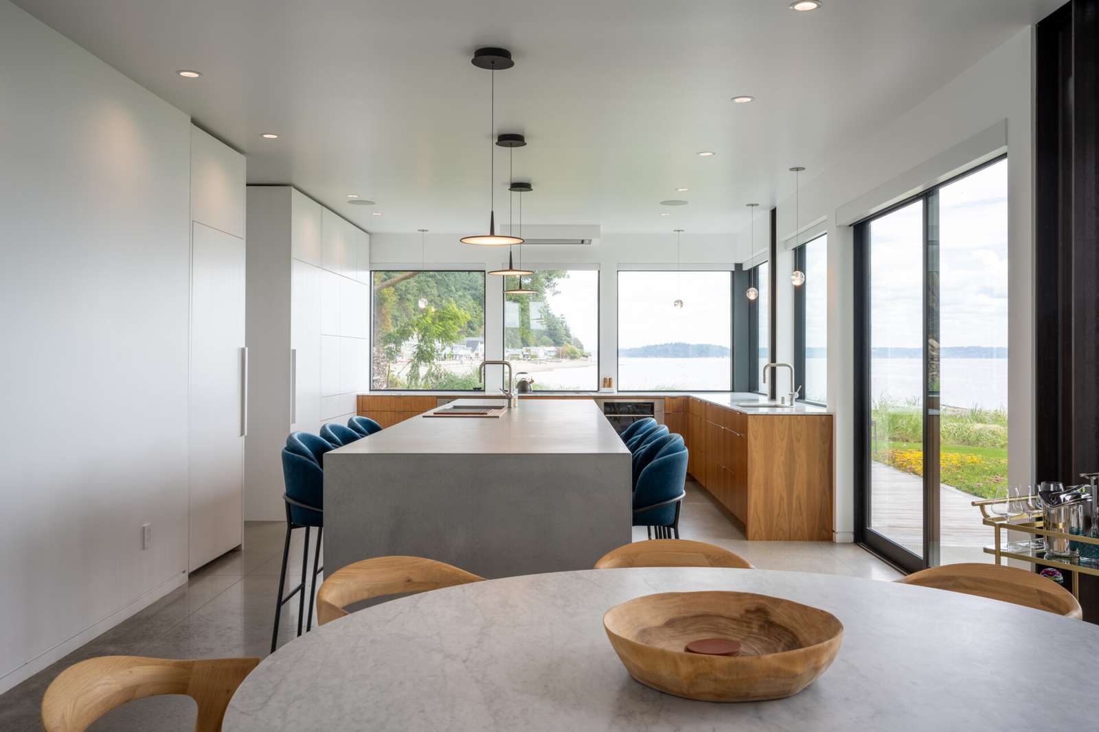 The kitchen has a long concrete island anchors the space, providing room for everyday use and casual gathering. Minimalist white cabinetry is paired with warm wood base units, keeping the palette restrained while adding subtle contrast. Floor to ceiling windows line the perimeter, drawing in natural light and uninterrupted water views.