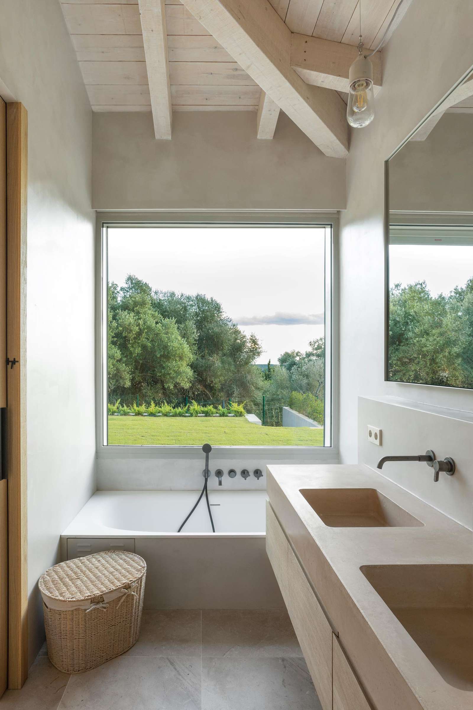 A modern bathroom with a built-in bathtub below a large window, and a wood vanity with a thick countertop that also has built-in sinks.