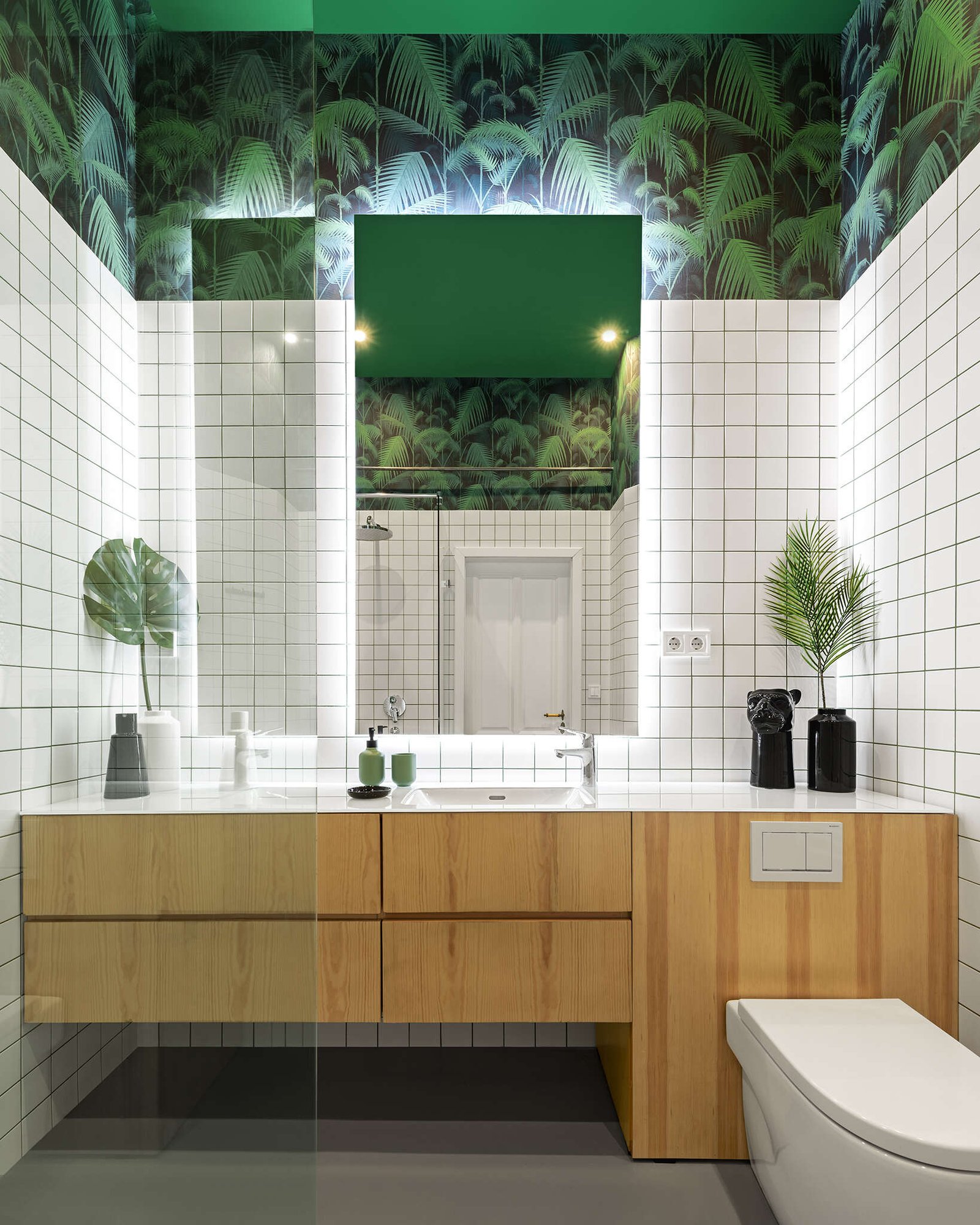 A clean and character filled bathroom with white tiles, a wood vanity, palm patterned walls, and softly back lit mirrors.