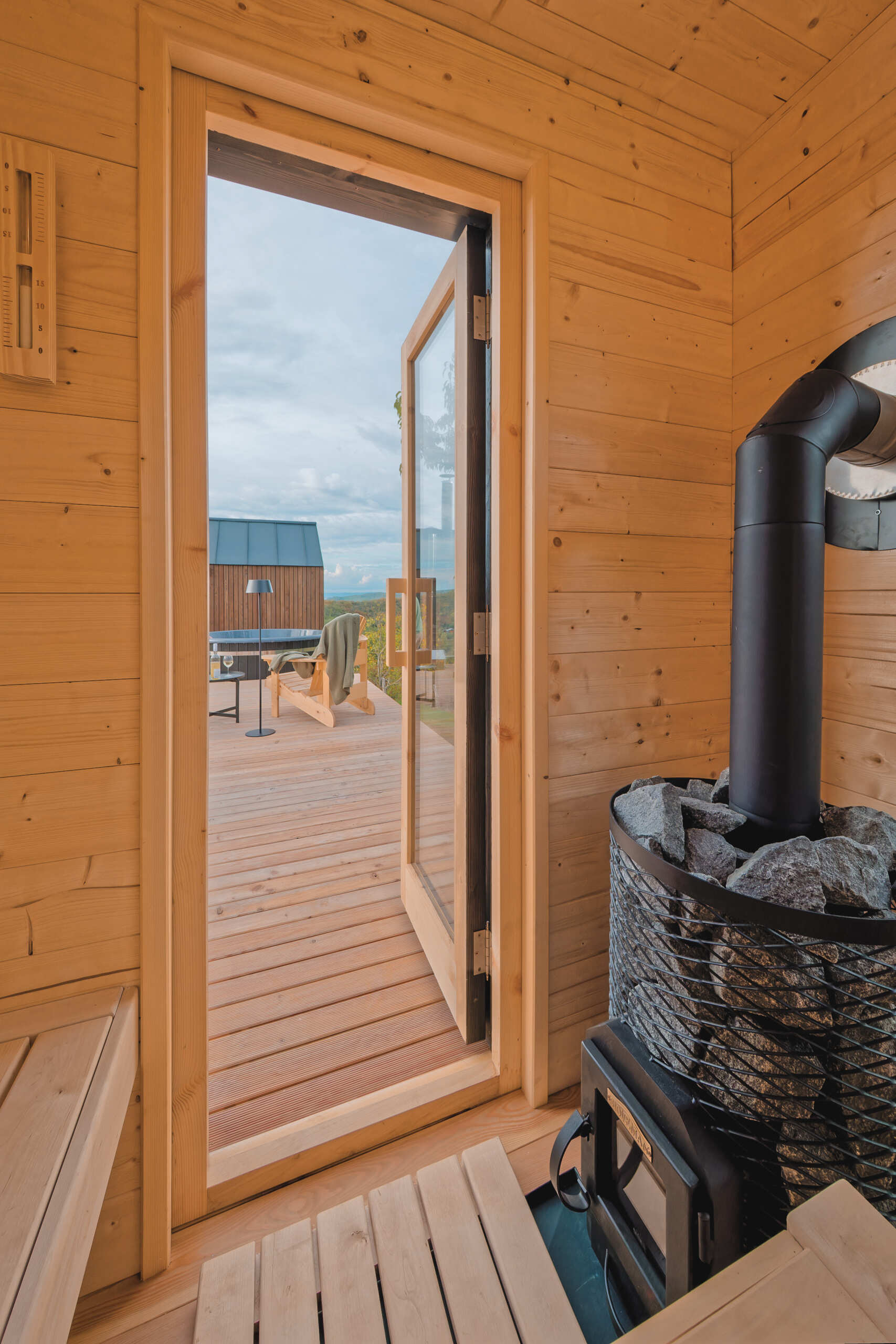 A large larch terrace extends the living space outdoors, leading to a hot tub and sauna that invite year-round use. Fire elements in the cabin, terrace, and sauna create warmth and atmosphere against the open hillside.