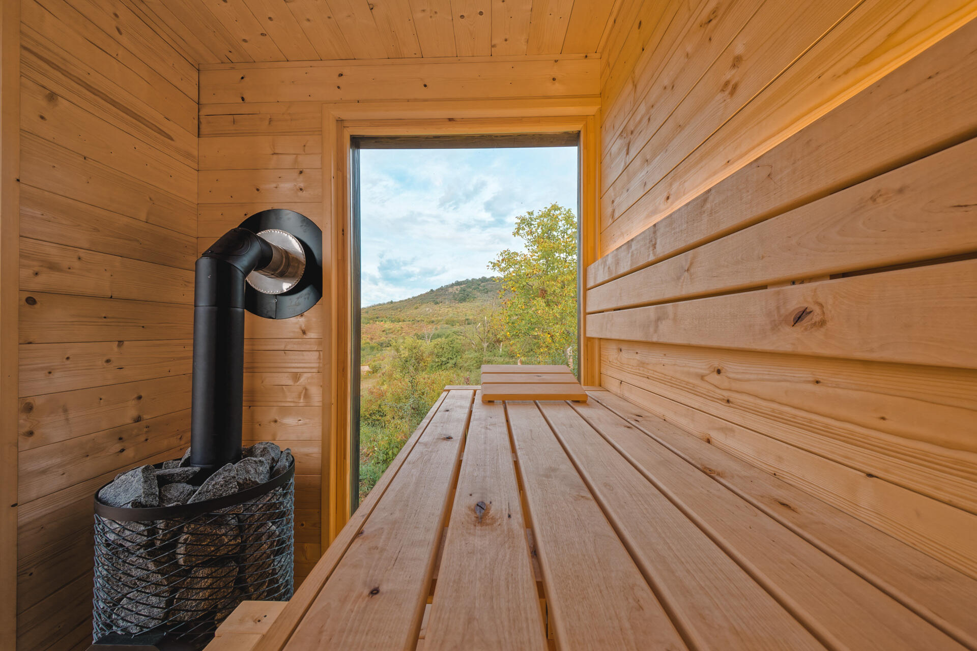 A large larch terrace extends the living space outdoors, leading to a hot tub and sauna that invite year-round use. Fire elements in the cabin, terrace, and sauna create warmth and atmosphere against the open hillside.
