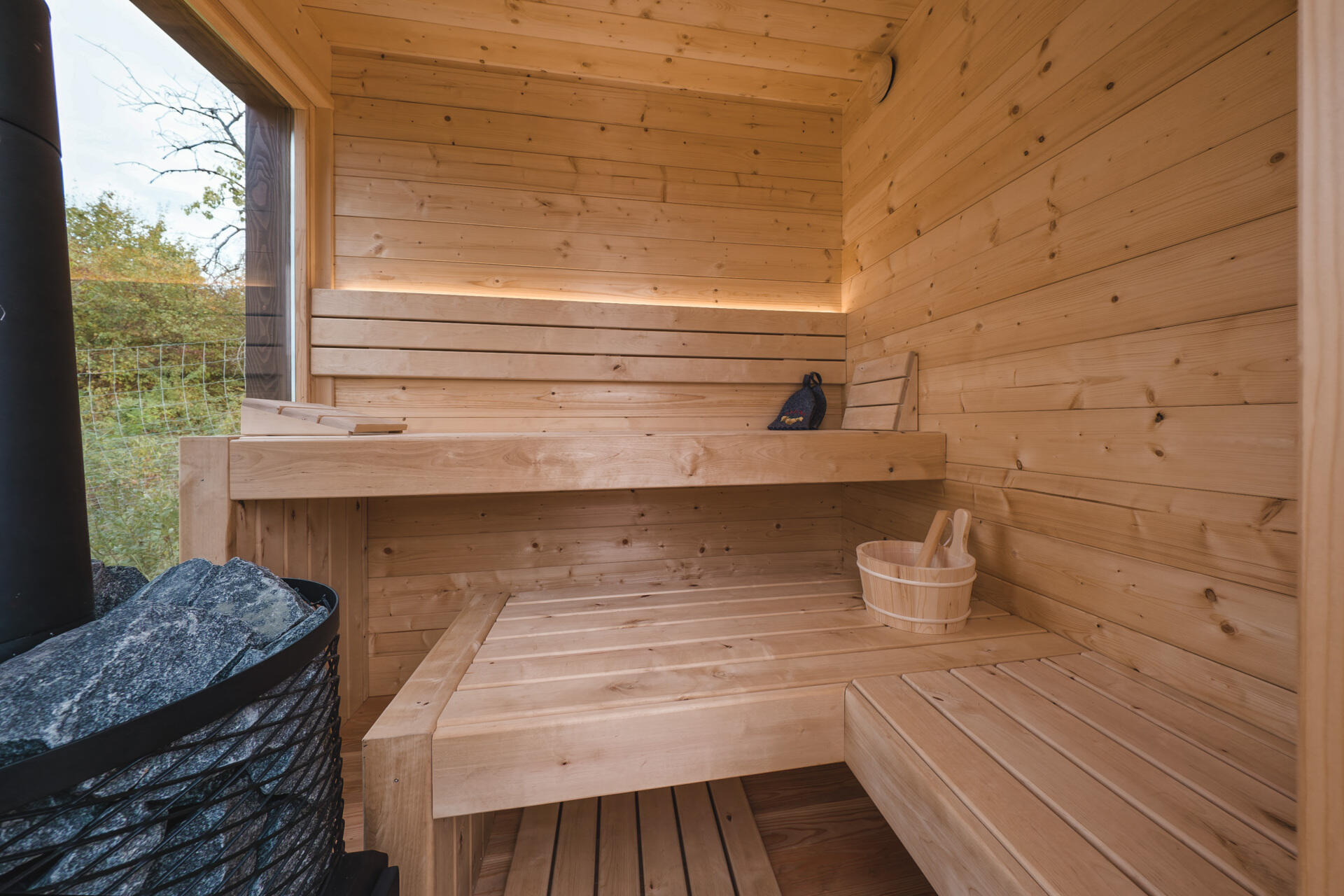 A large larch terrace extends the living space outdoors, leading to a hot tub and sauna that invite year-round use. Fire elements in the cabin, terrace, and sauna create warmth and atmosphere against the open hillside.