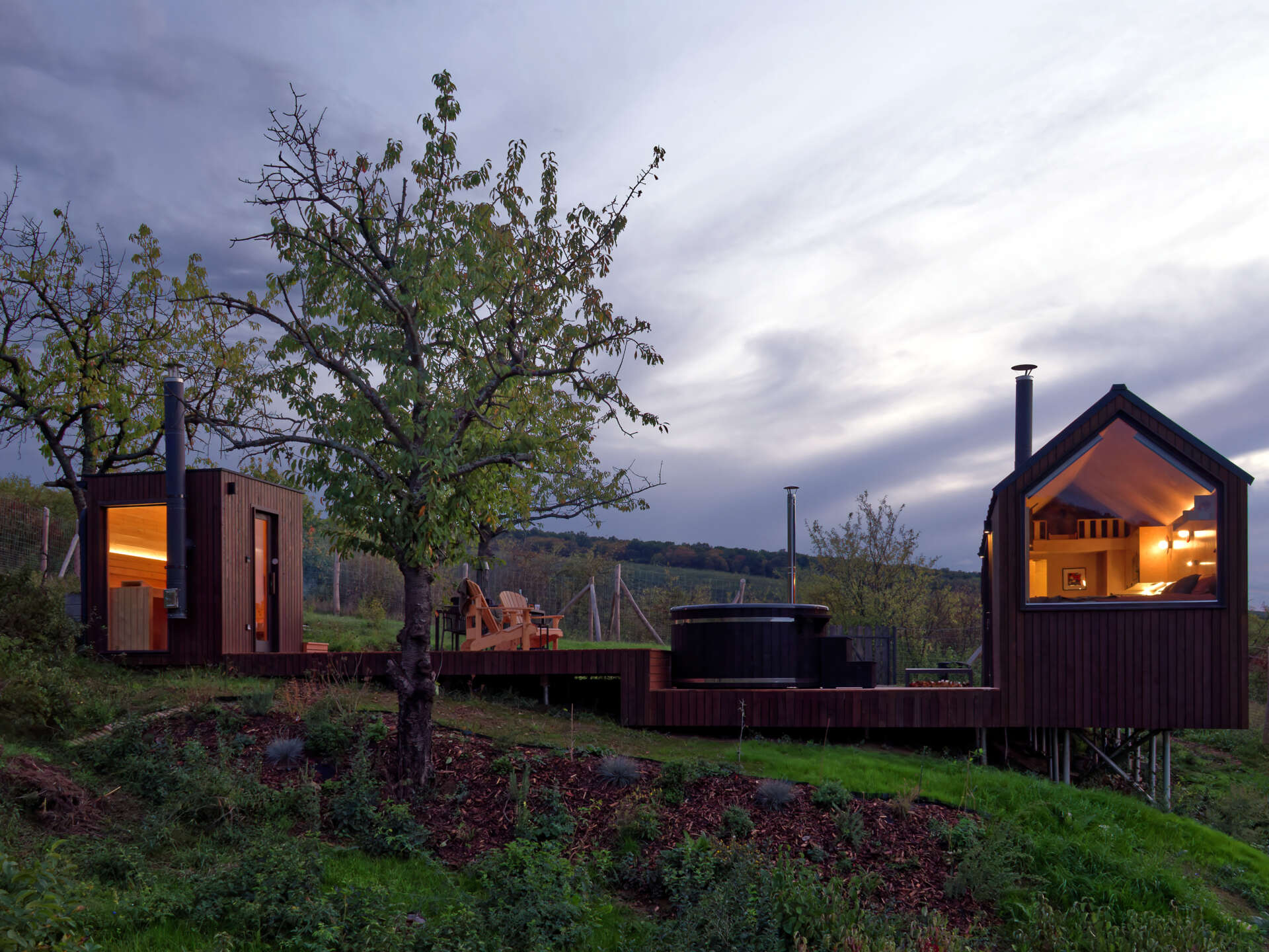 As night falls, the cabin glows softly above the village below. The illuminated trapezoidal window turns this small structure into a quiet landmark in the Hungarian hills, showing how thoughtful design transforms even the simplest form.