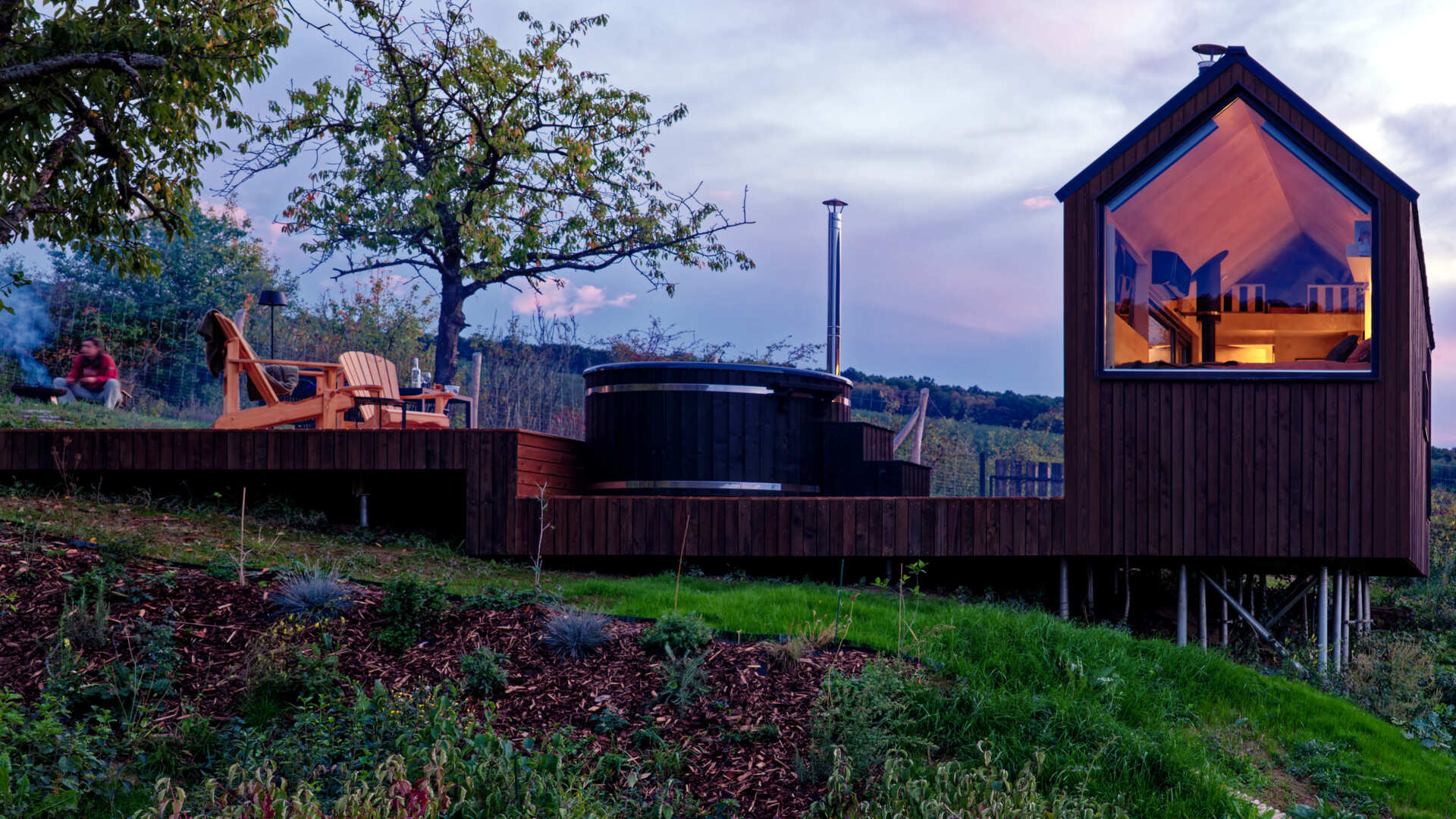 As night falls, the cabin glows softly above the village below. The illuminated trapezoidal window turns this small structure into a quiet landmark in the Hungarian hills, showing how thoughtful design transforms even the simplest form.