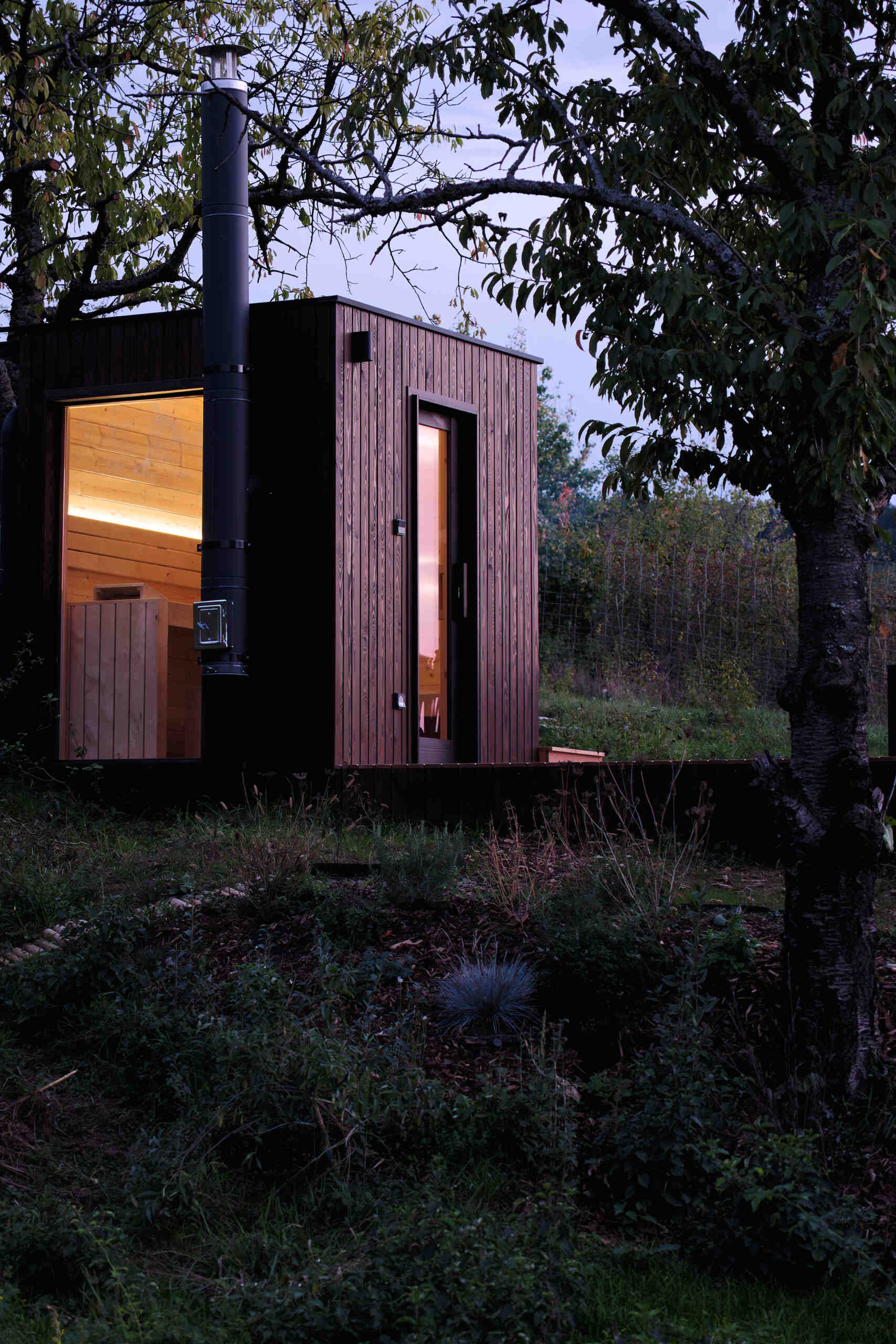 As night falls, the cabin glows softly above the village below. The illuminated trapezoidal window turns this small structure into a quiet landmark in the Hungarian hills, showing how thoughtful design transforms even the simplest form.