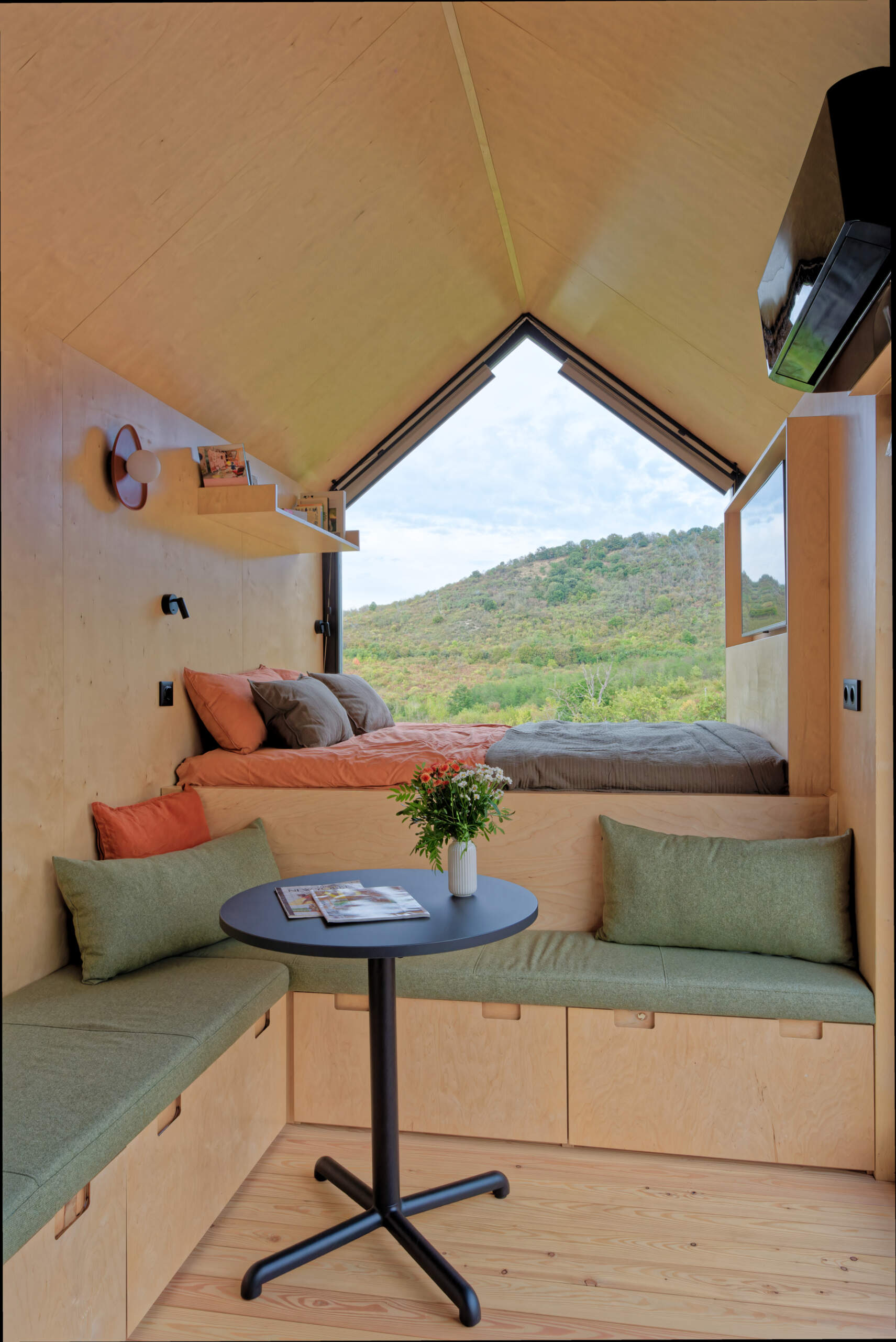 Light birch plywood, a visible gabled roof, and a striking trapezoidal window make this micro cabin interior feel surprisingly spacious. Included in the cabin is a sleeping area, lounge, kitchen, loft, and bathroom.