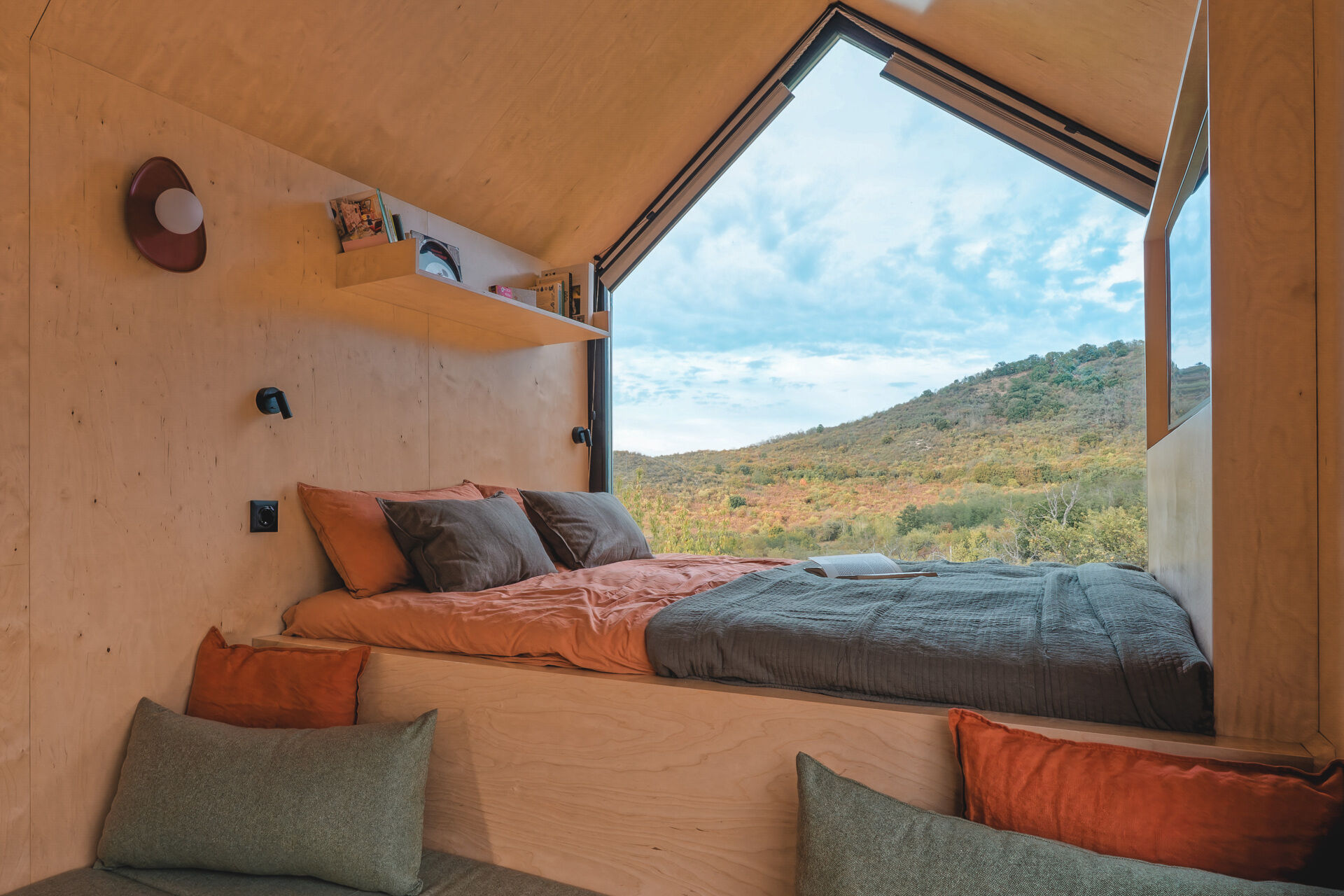Light birch plywood, a visible gabled roof, and a striking trapezoidal window make this micro cabin interior feel surprisingly spacious. Included in the cabin is a sleeping area, lounge, kitchen, loft, and bathroom.