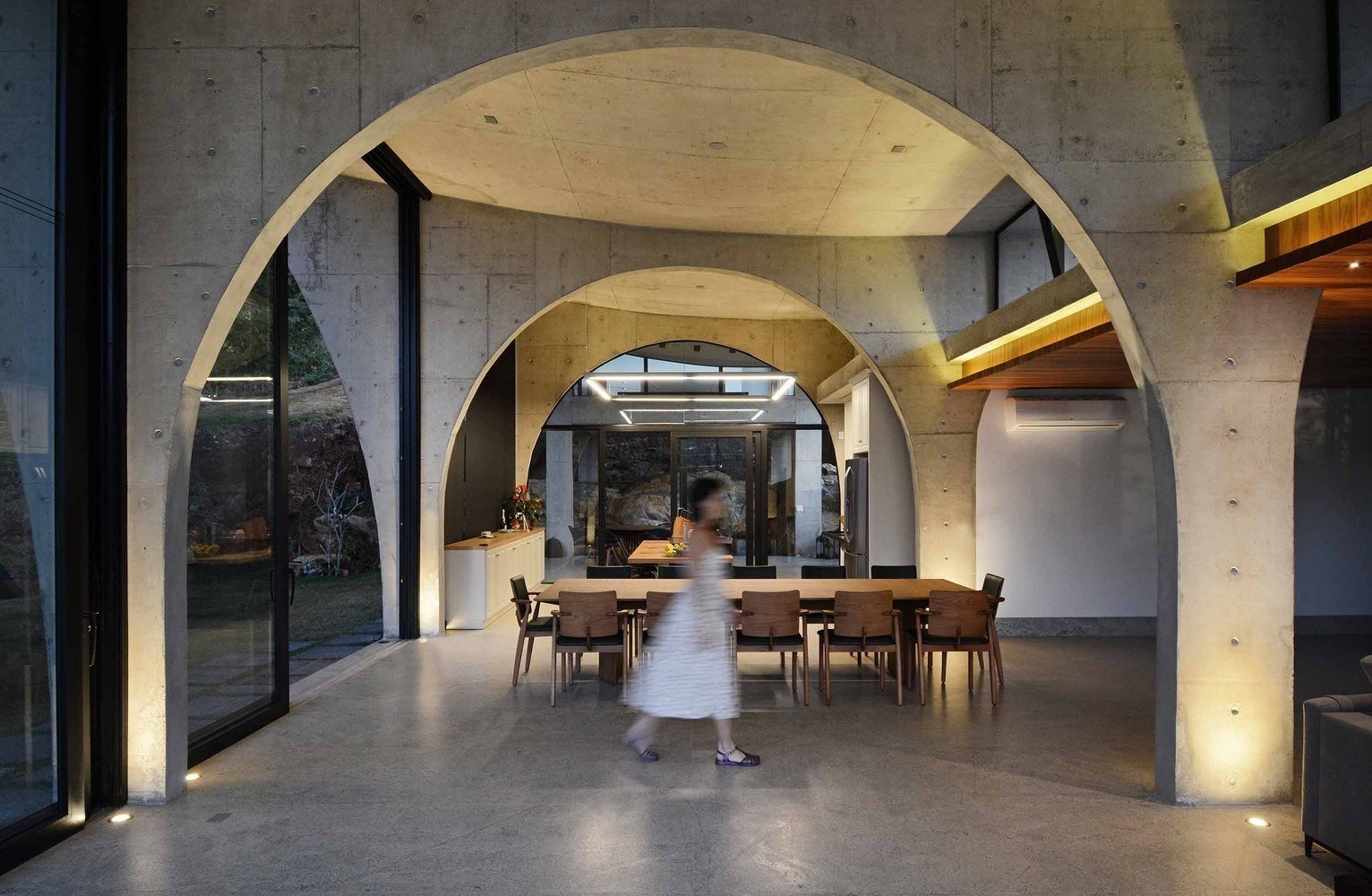 The living room, dining area, and kitchen share a dramatic double height space framed by concrete arches and large sliding glass doors that connect the interior to the gardens.