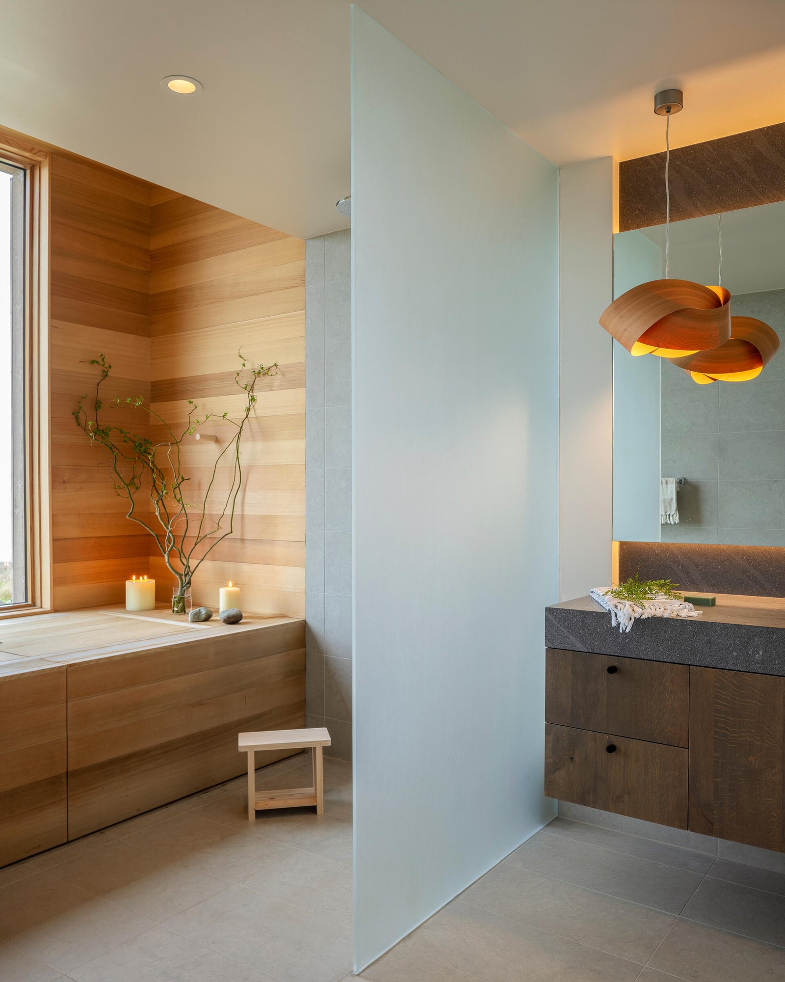 The bathroom continues the same material palette found throughout the home, with a built-in soaking tub hidden beneath a wood cover and positioned beneath the window.