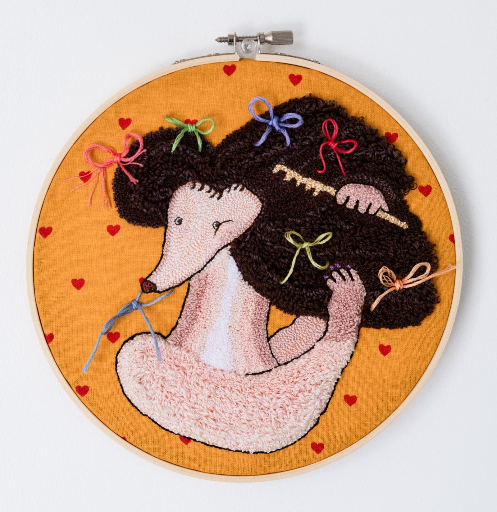 an embroidery by Misato Sano depicting a dog combing its hair, wearing colorful bows. the piece sits inside an embroidery hoop