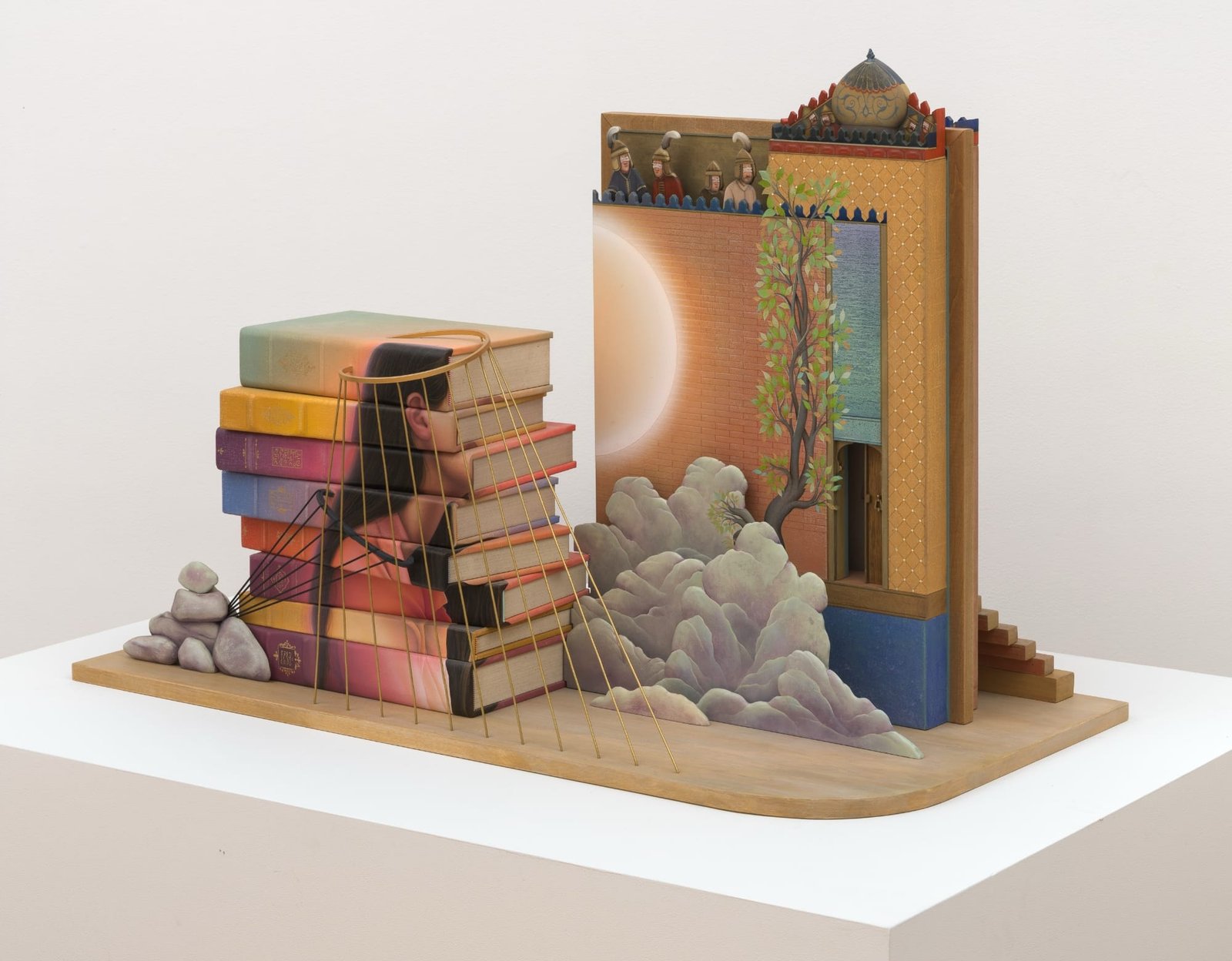 a sculpture with a stack of books, clouds, a building, and a woman's silhouette bound with gold cords