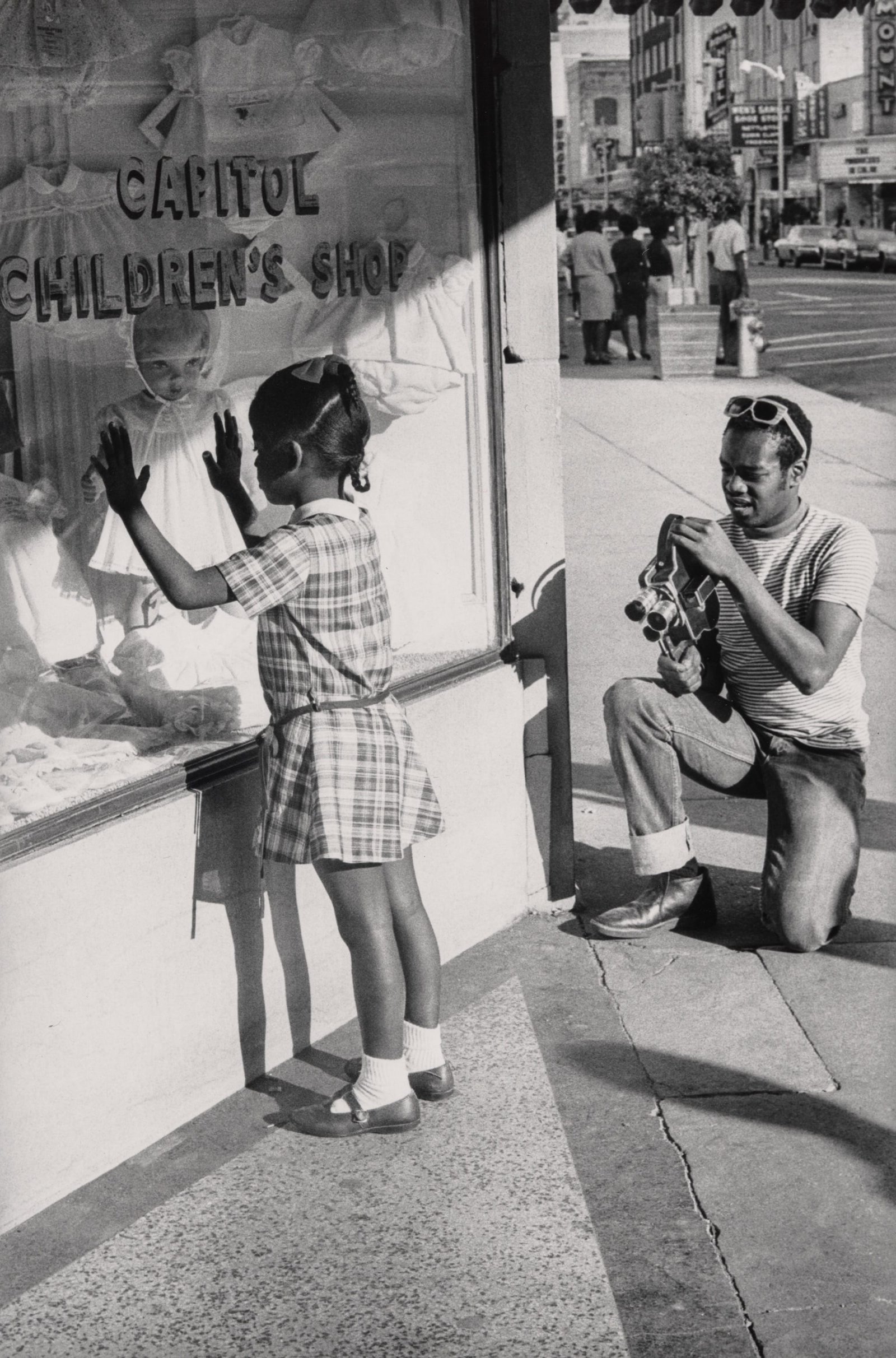 a black and white photo of a photographer capturing a young Black girl looking into a shop window
