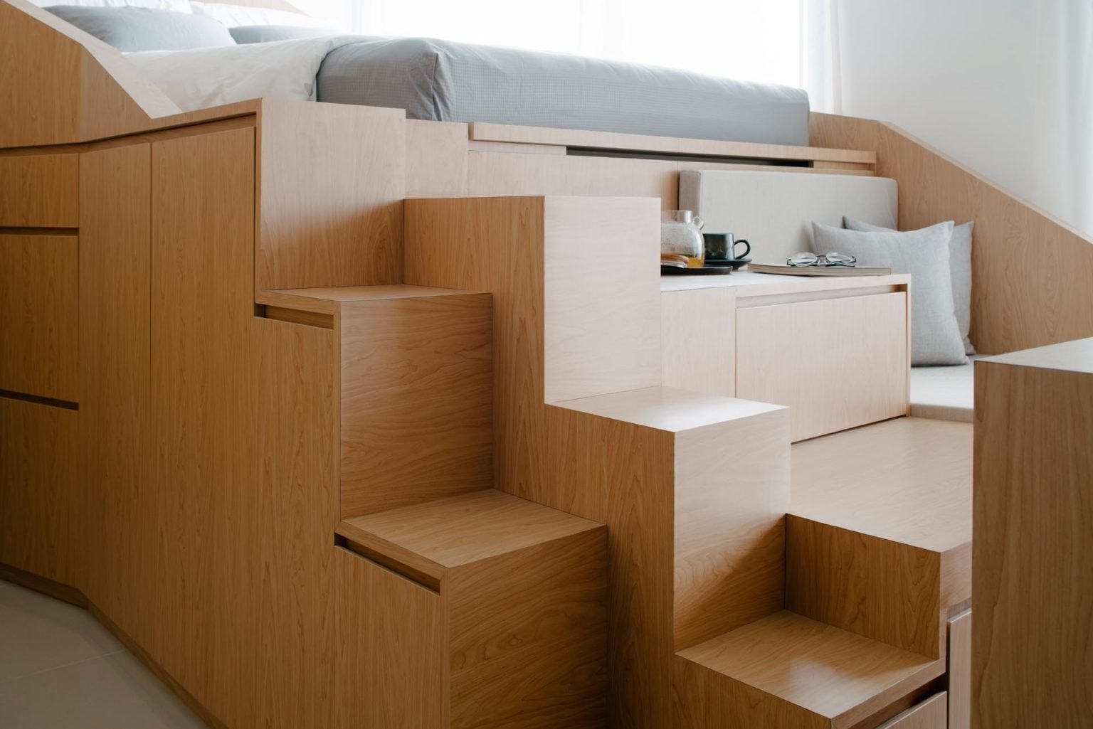 This custom wood platform combines a loft bed, sofa, dining table, workspace, stairs, and storage into one smart design for compact apartment living.