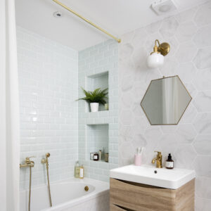 الحمام بجدران وأرضيات بيضاء وبانيو ومرآة ذهبية فوق وحدة زينة خشبية.