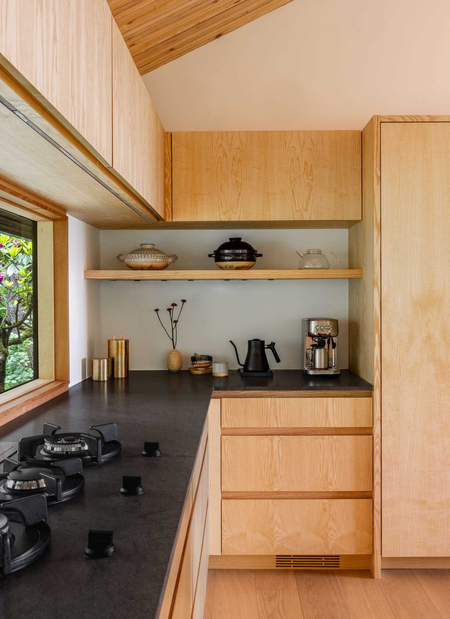 The kitchen includes a horizontal window above the cooktop, offering views of mature rhododendrons, as well as plain-sawn Ash cabinetry and Basaltina Natural Stone countertop with a honed finish.