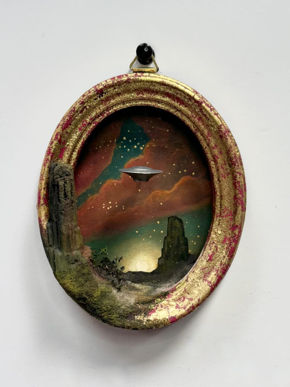 within a miniature ovoid frame, a detailed diorama depicts a landscape with a hovering UFO