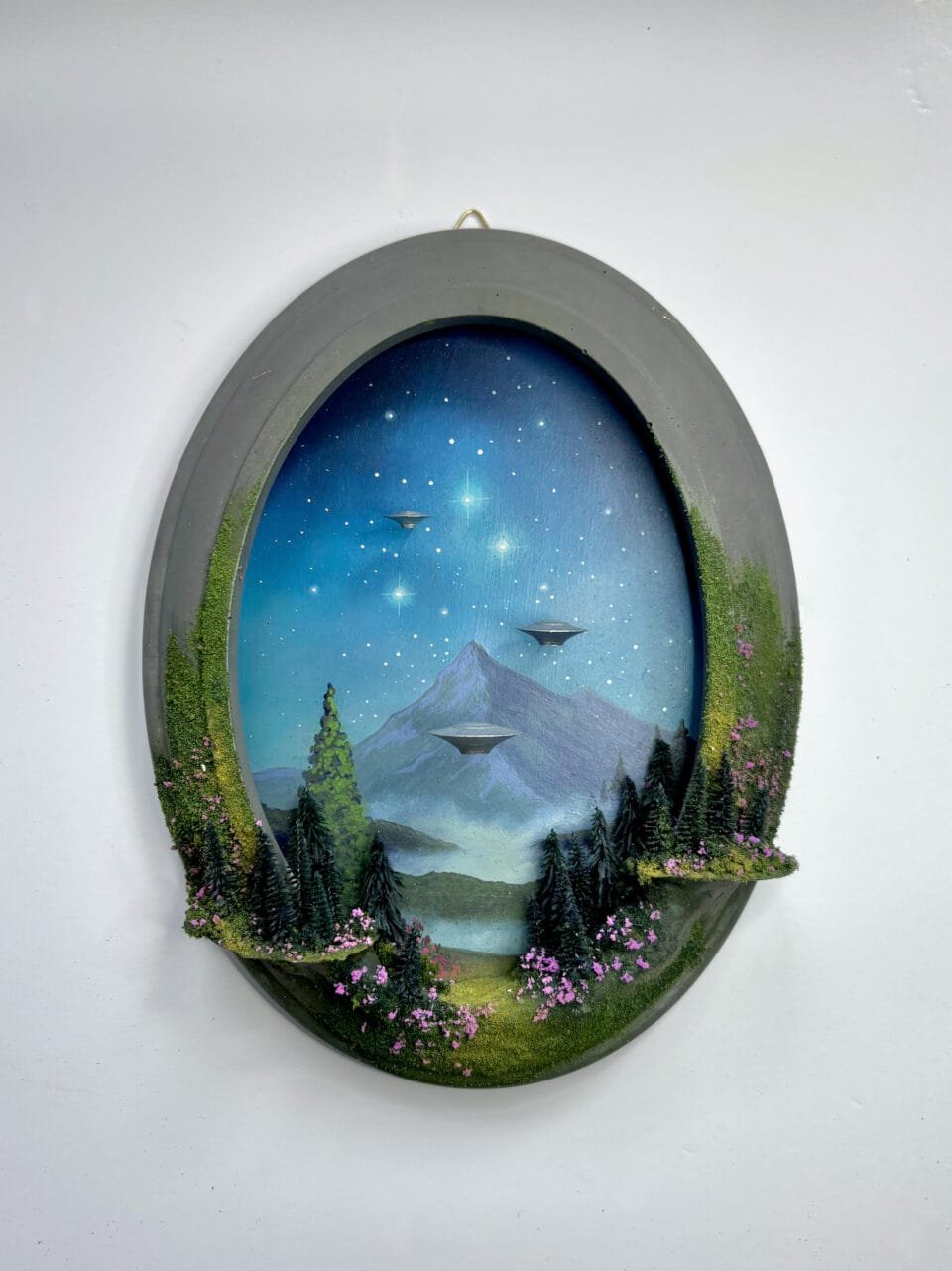 within a miniature ovoid frame, a detailed diorama depicts a landscape with hovering UFOs