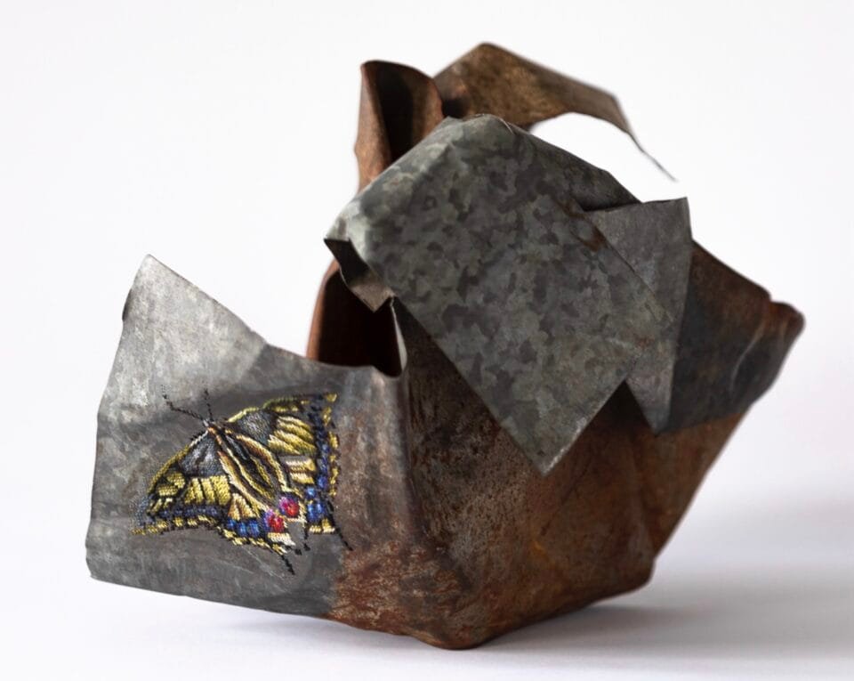 a rusted, crumpled ball of metal with a cross-stitched butterfly in one part