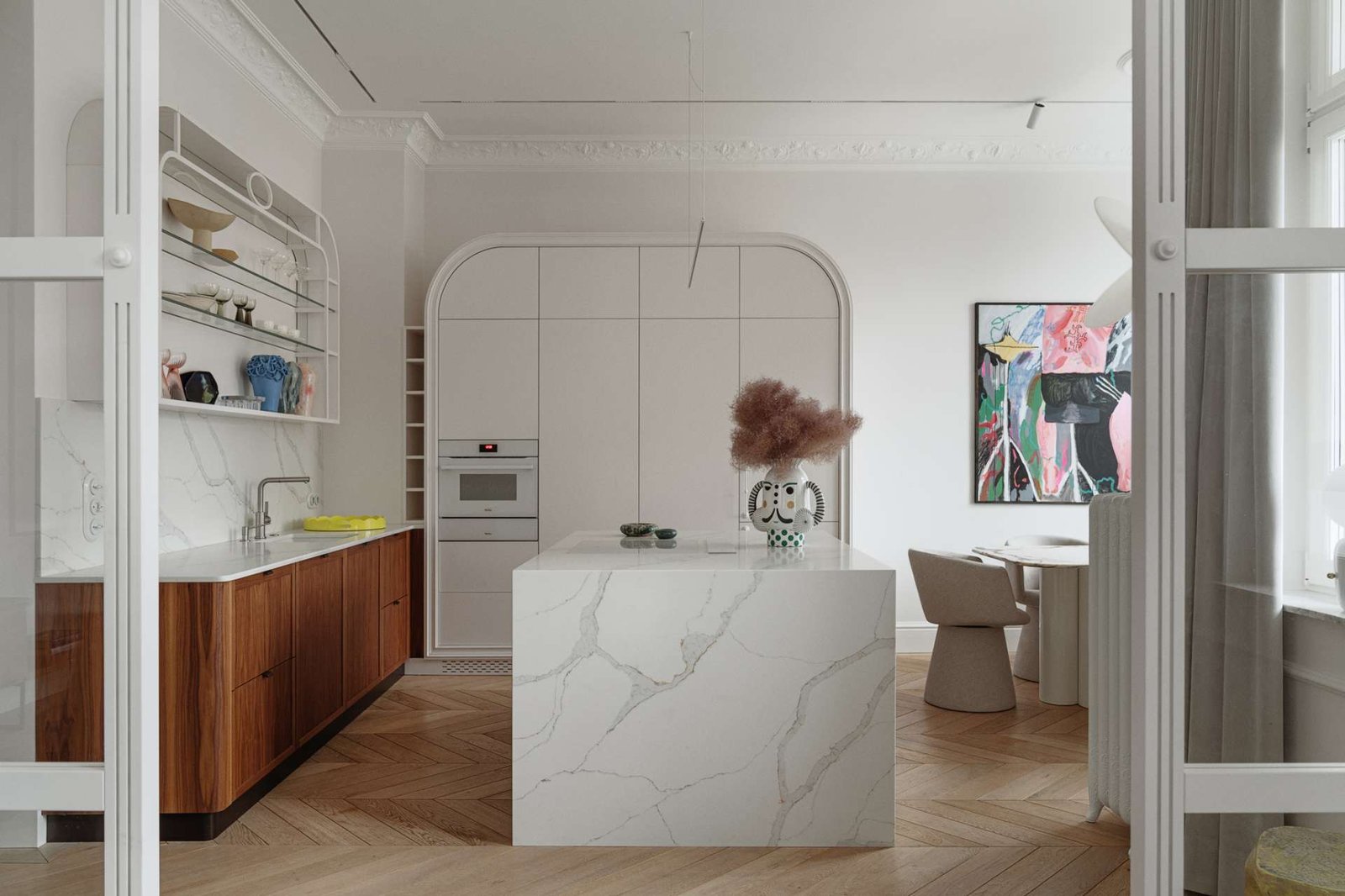 This kitchen pairs sculptural softness with practical sophistication. A central island in marble-look stone anchors the space, its clean lines echoed in the curved cabinetry and arched built-ins. Walnut lower cabinets add warmth and grain, while open shelving displays a curated mix of ceramics and glassware.