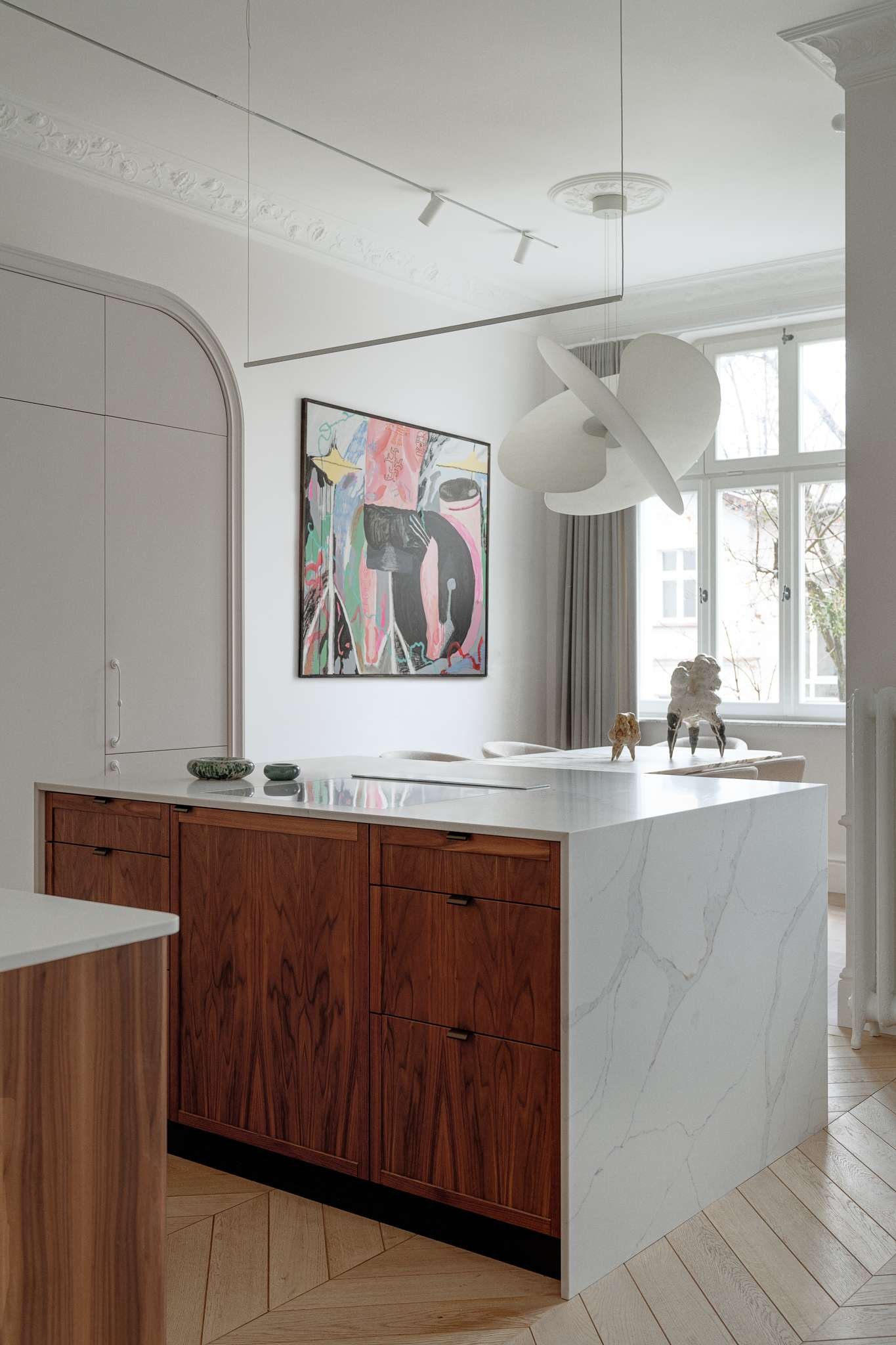 This kitchen pairs sculptural softness with practical sophistication. A central island in marble-look stone anchors the space, its clean lines echoed in the curved cabinetry and arched built-ins. Walnut lower cabinets add warmth and grain, while open shelving displays a curated mix of ceramics and glassware.