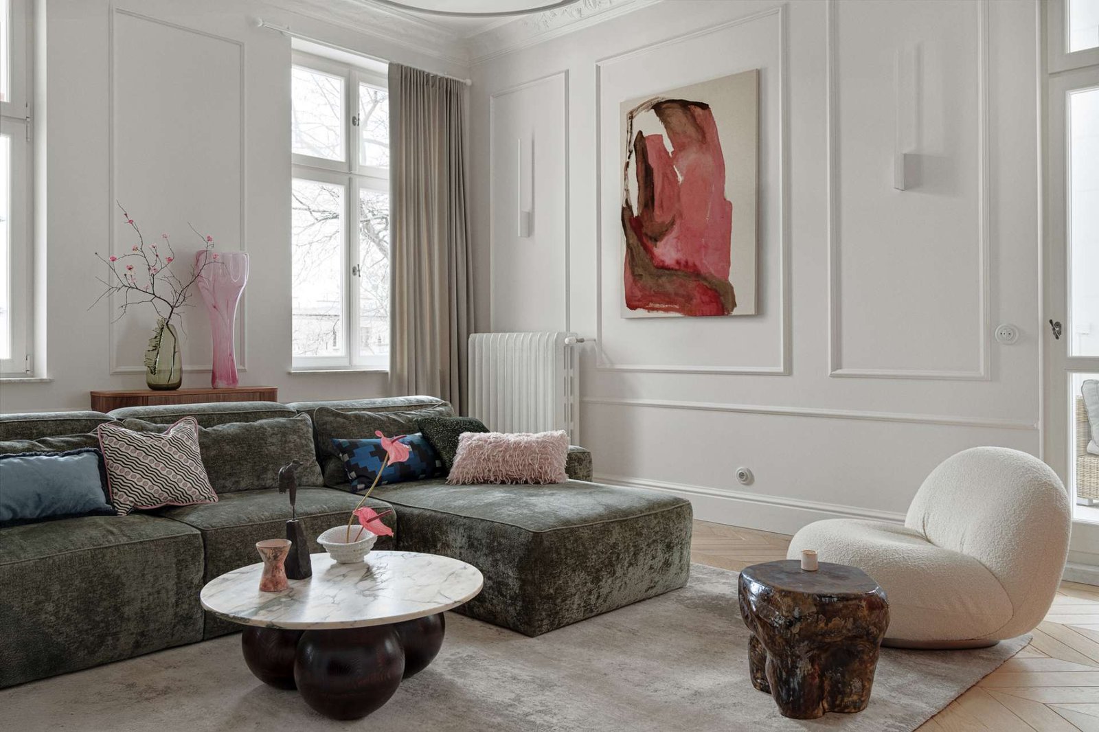 This living room combines refined elegance with cozy textures and bold accents. A plush green sectional anchors the space, softened by layered cushions in playful prints and hues. The marble-topped coffee table rests on rounded spheres, grounding the room with sculptural weight. A curved white lounge chair and a deep-toned side table add contrast and shape, while a vibrant pink abstract painting by Aleksandra Szewczuk brings warmth to the pale walls.