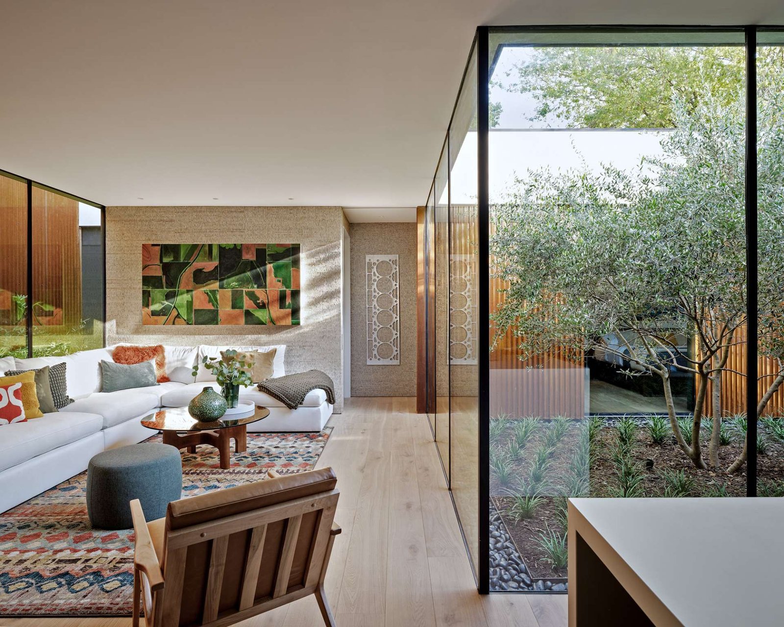 This cozy living room blends earthy textures and warm wood tones with expansive glass walls that open to lush bamboo landscaping. A colorful rug and sculptural furnishings add comfort and character to the modern minimalist space.
