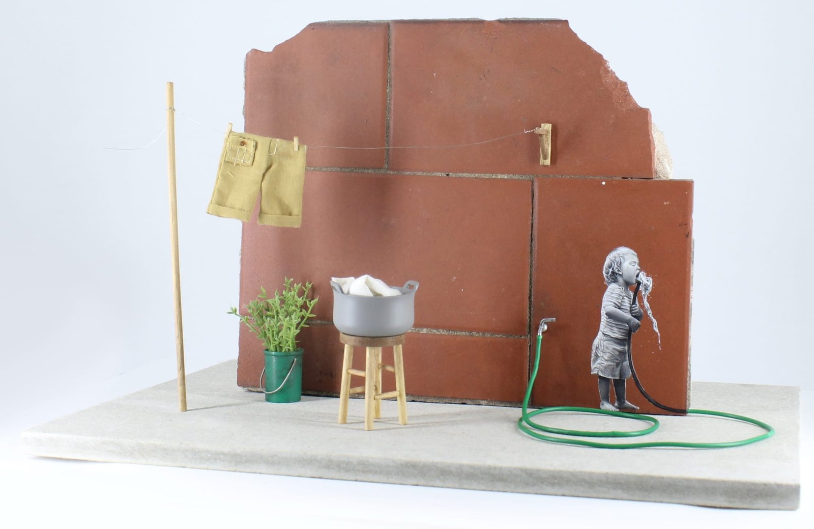 a child drinking from a green hose painted on tile with a wash bucket and laundry on the line nearby