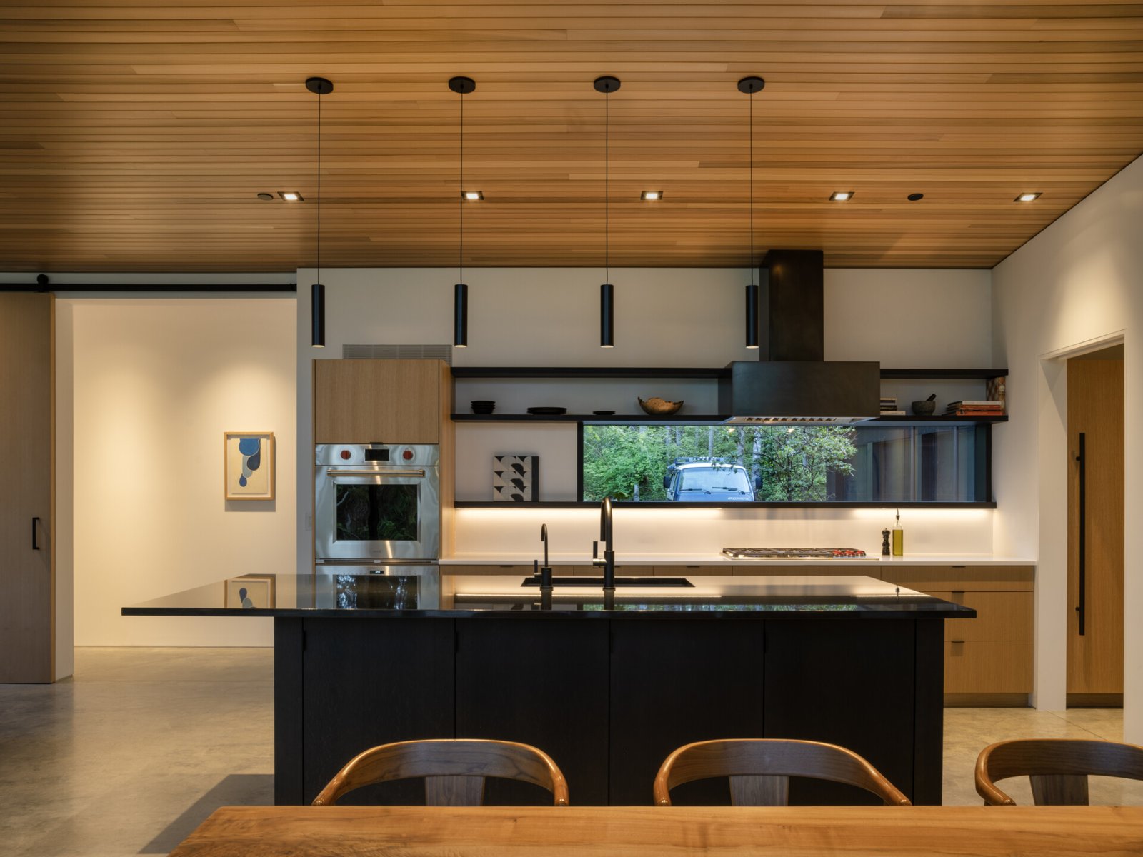 This modern kitchen combines modern clarity with natural warmth. A wood-paneled ceiling adds texture above a streamlined black island and matte fixtures. Open shelving and minimalist cabinetry offer practical storage while keeping the space visually quiet and functional for daily use.