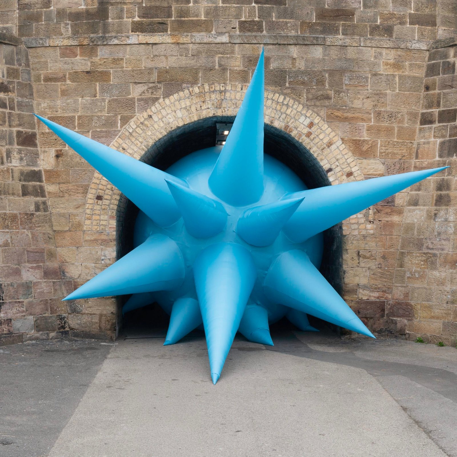 a large-scale installation by Steve Messam under an old stone bridge of a blue, spiked form that appears wedged in the archway under the bridge